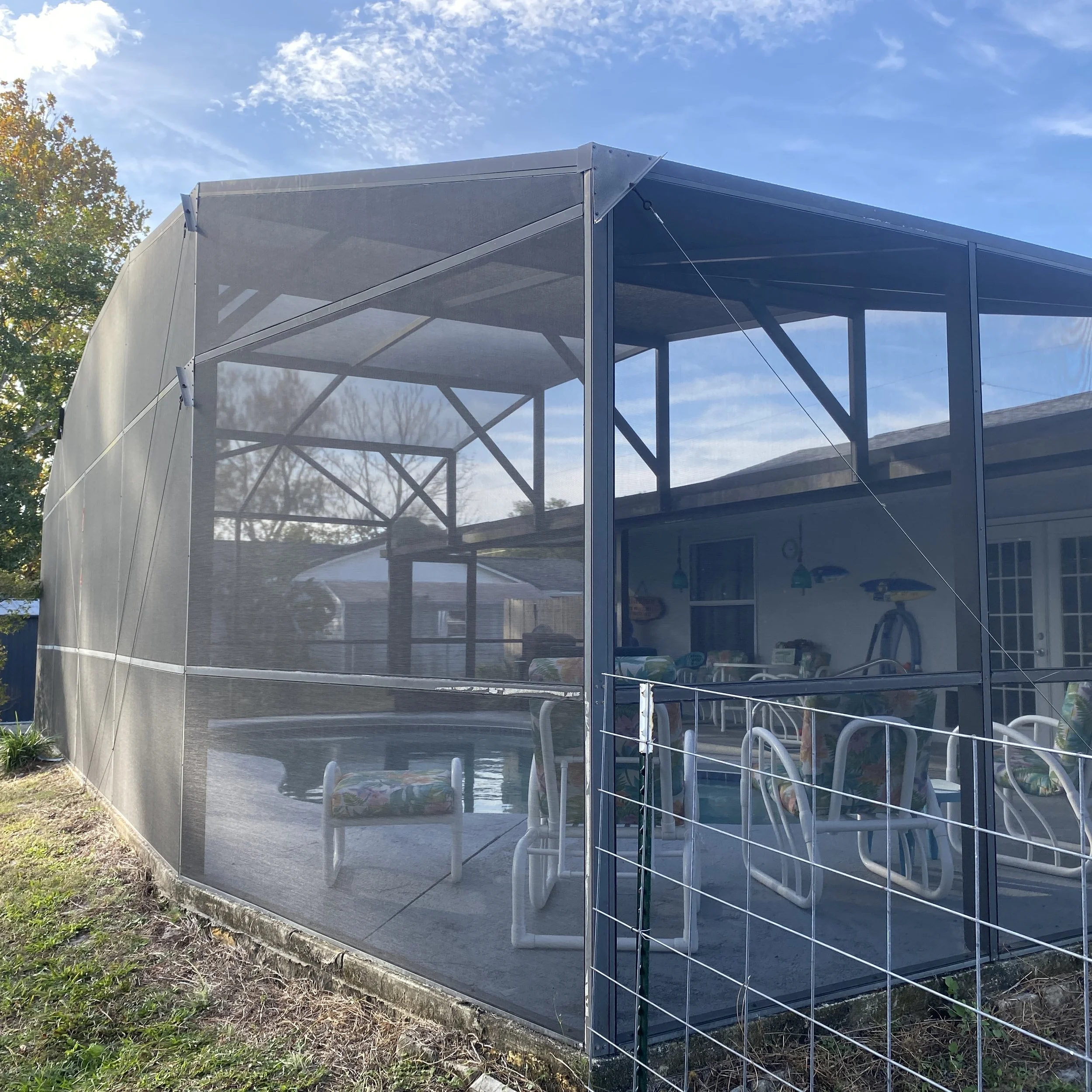 Screened-in backyard patio with outdoor furniture, pool, and climbing equipment, enclosed by a mesh screen with supporting wires, in a suburban backyard under a blue sky.