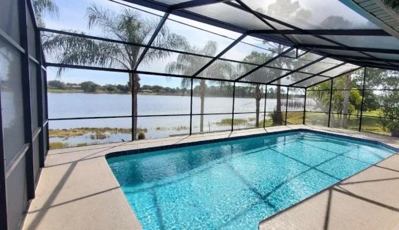 Swimming pool enclosed by a screen with a view of a lake and palm trees in the background.