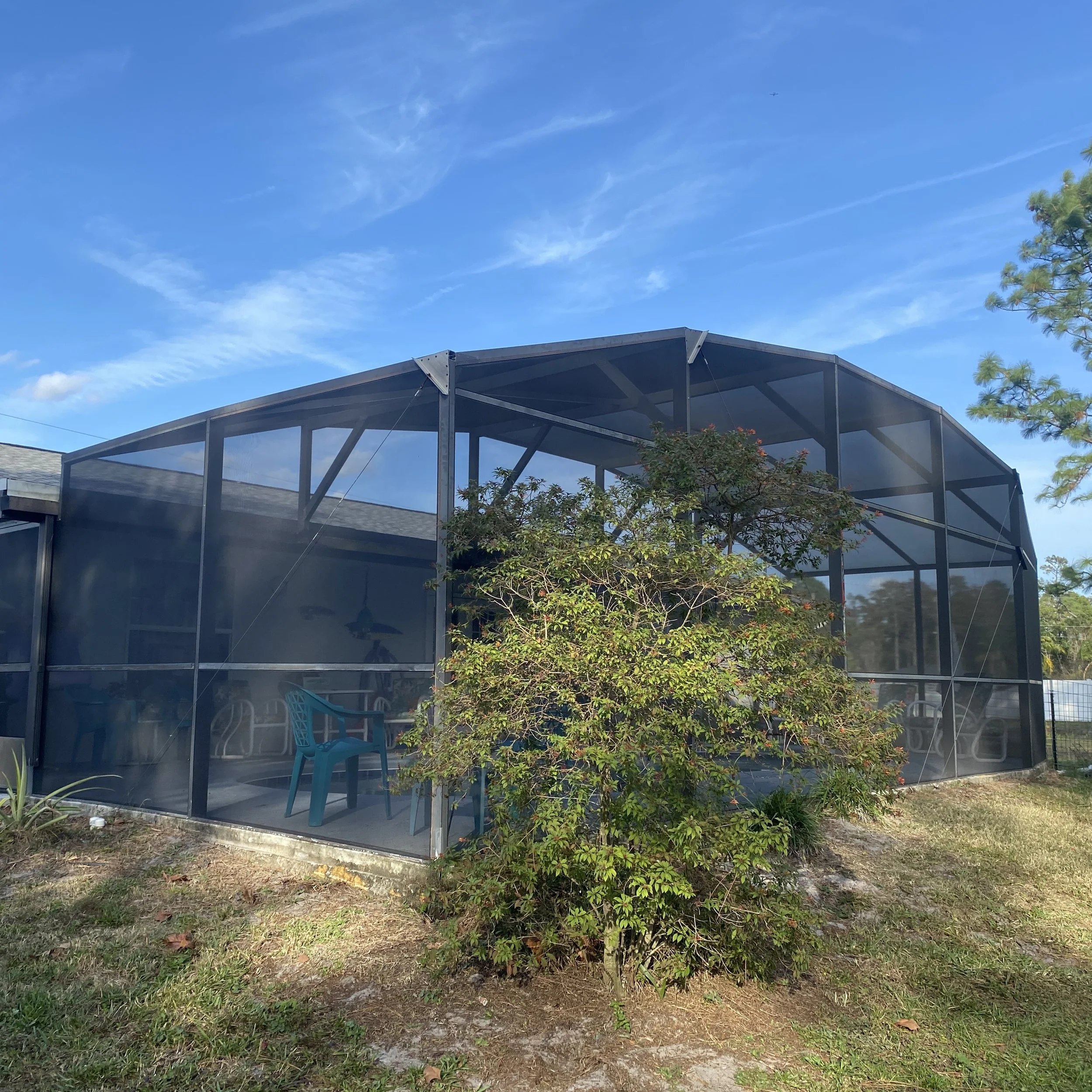 Screened porch with outdoor furniture and a small bush in front, under a blue sky with some clouds.