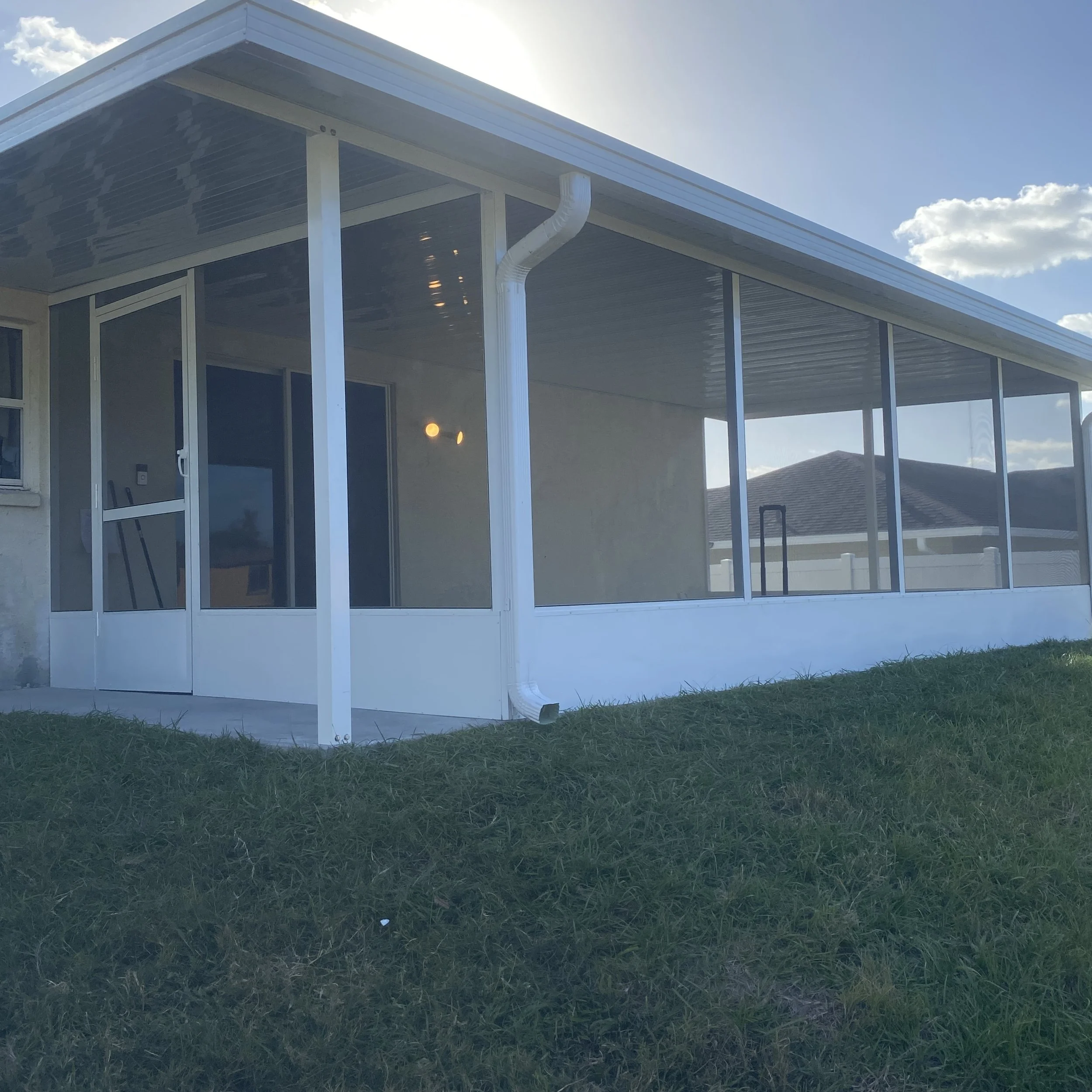 A screened porch attached to a house with white framing, a door, and a roof overhang, overlooking a grassy yard.