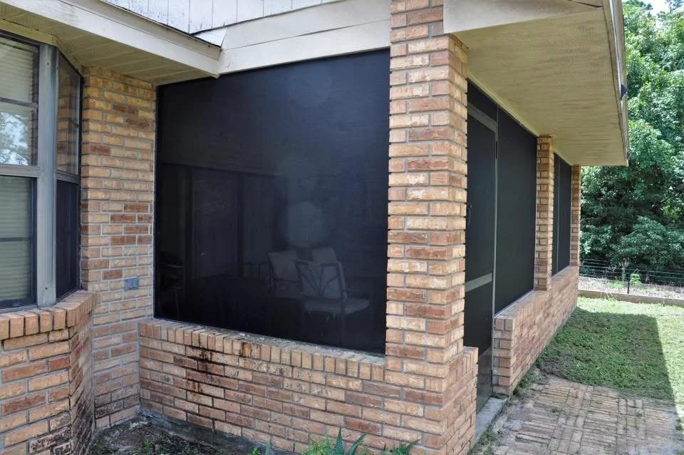 View of a brick house with a screened-in porch, black screening material, and a small garden area outside.