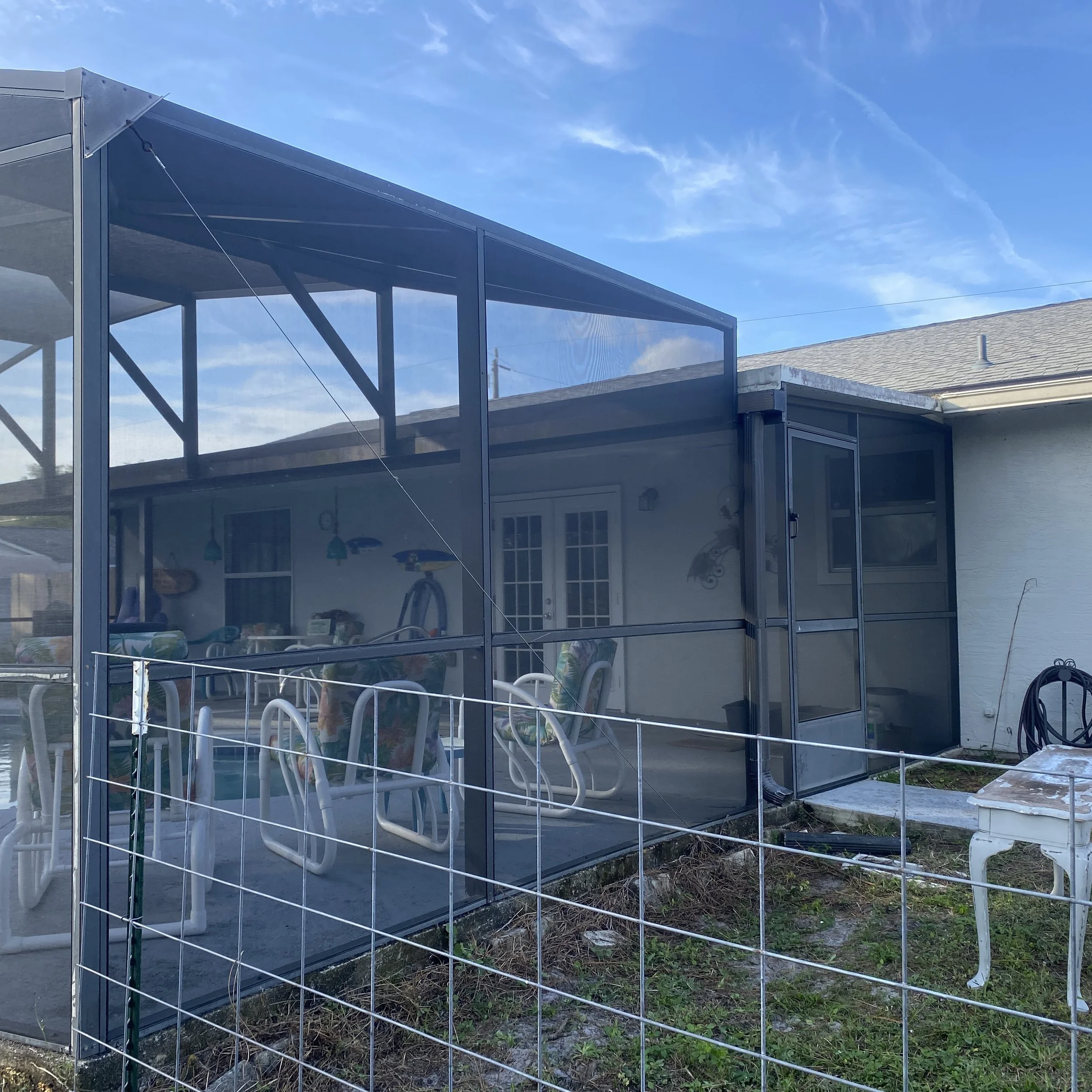 A screened porch attached to a house with outdoor furniture, including chairs with floral cushions, visible through the mesh screen enclosure on a sunny day.