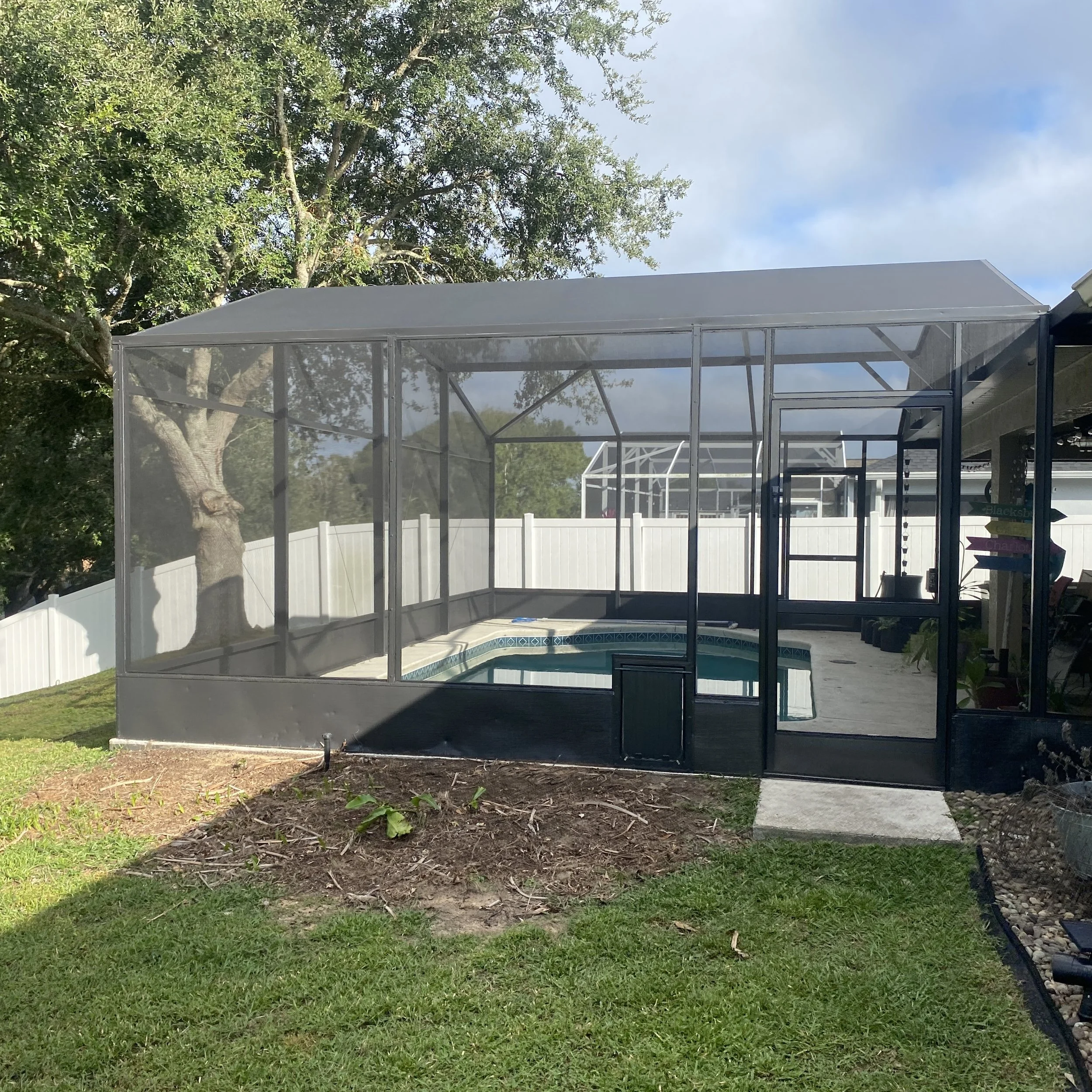  screened-in pool enclosure with a swimming pool inside, adjacent to a patio area and a white fence, with trees in the background.