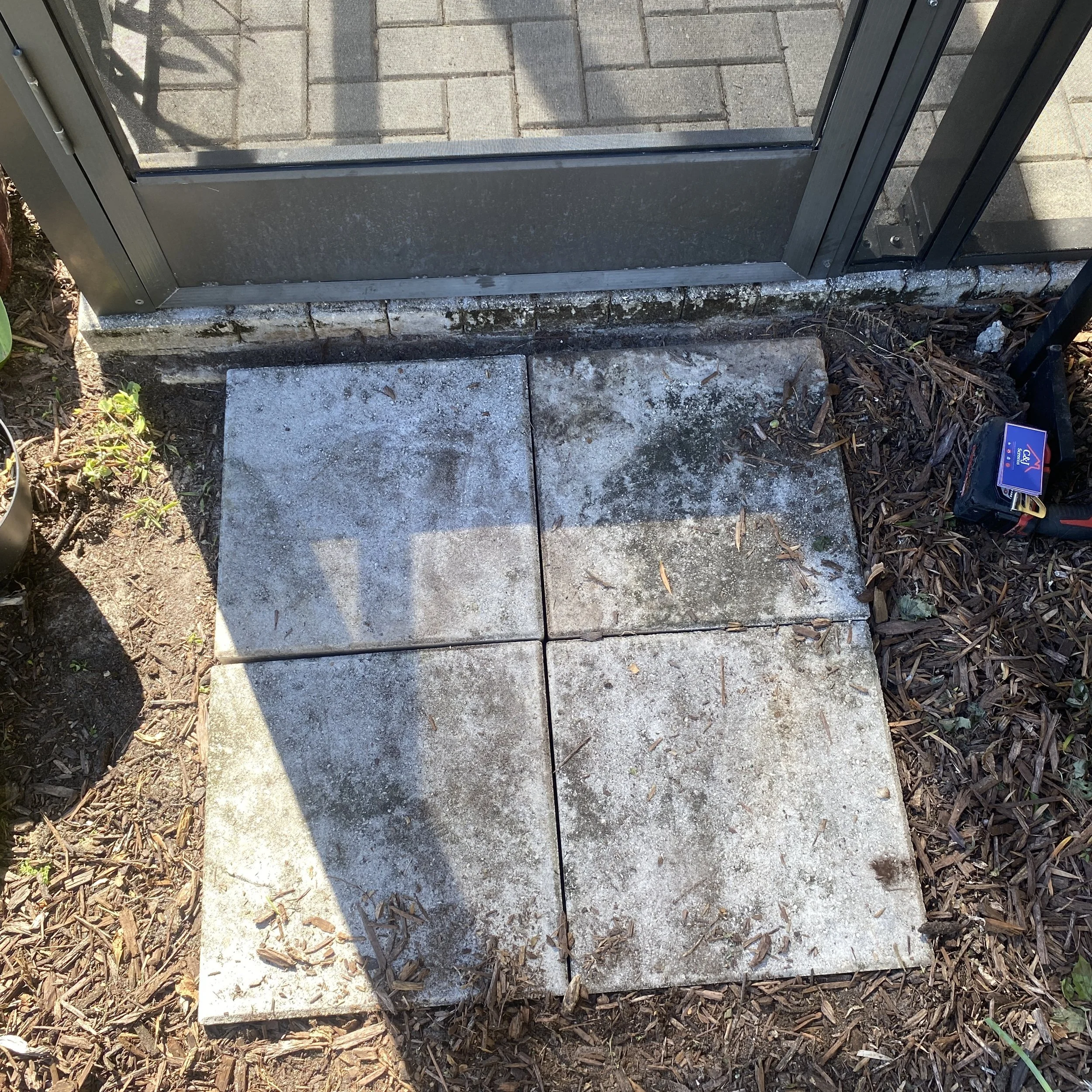 Four concrete tiles forming a small outdoor patio area, adjacent to a glass door and surrounded by soil and mulch.