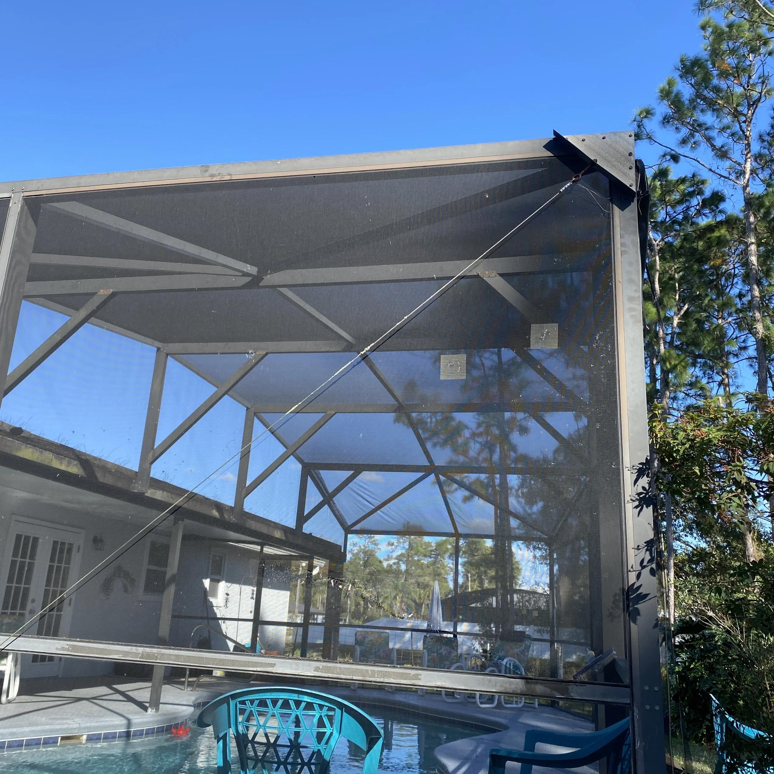 View of an enclosed porch area with a screened barrier, a swimming pool, outdoor furniture, and trees in the background under a clear blue sky.