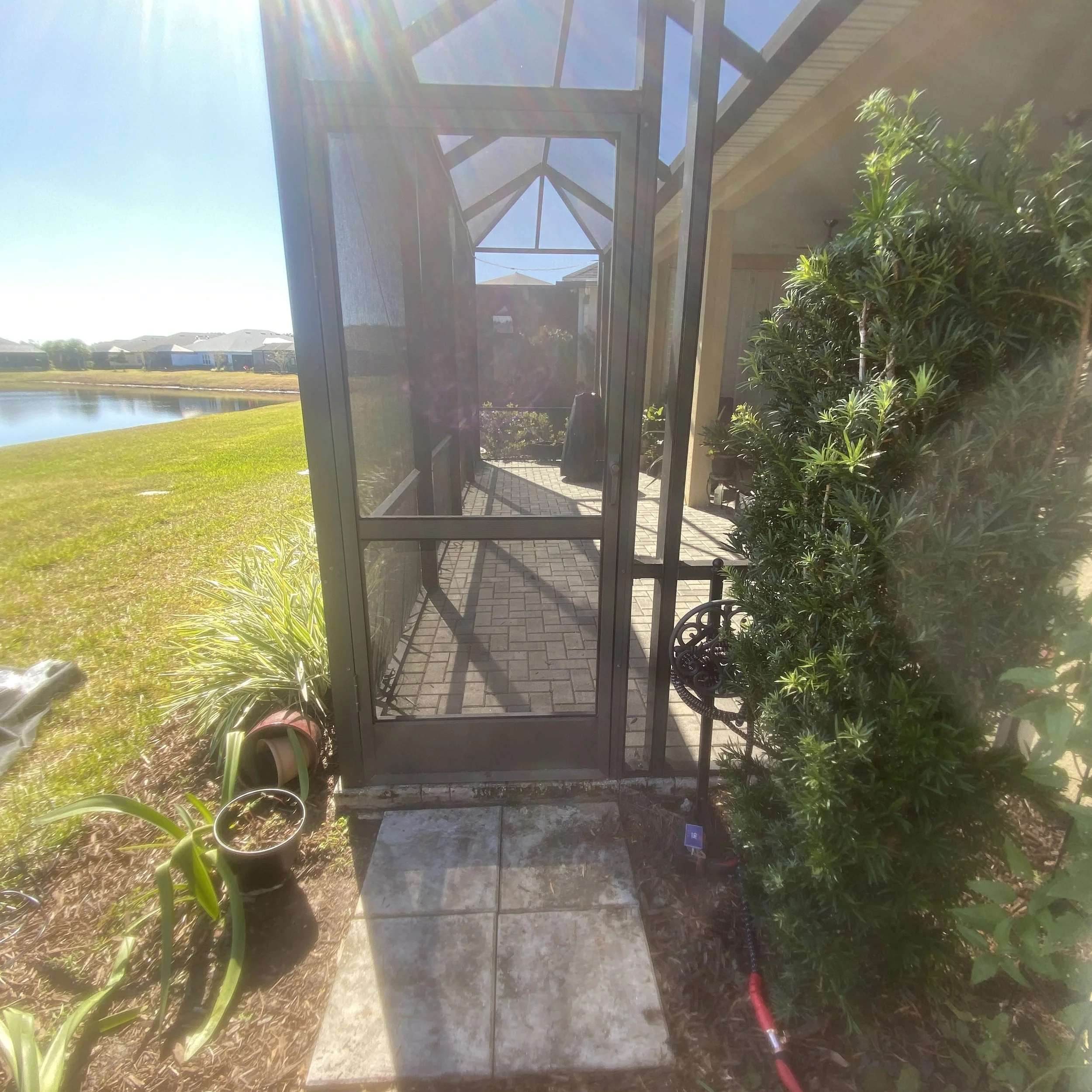 View of a screened porch with brick flooring, surrounded by plants, beside a pond on a sunny day.