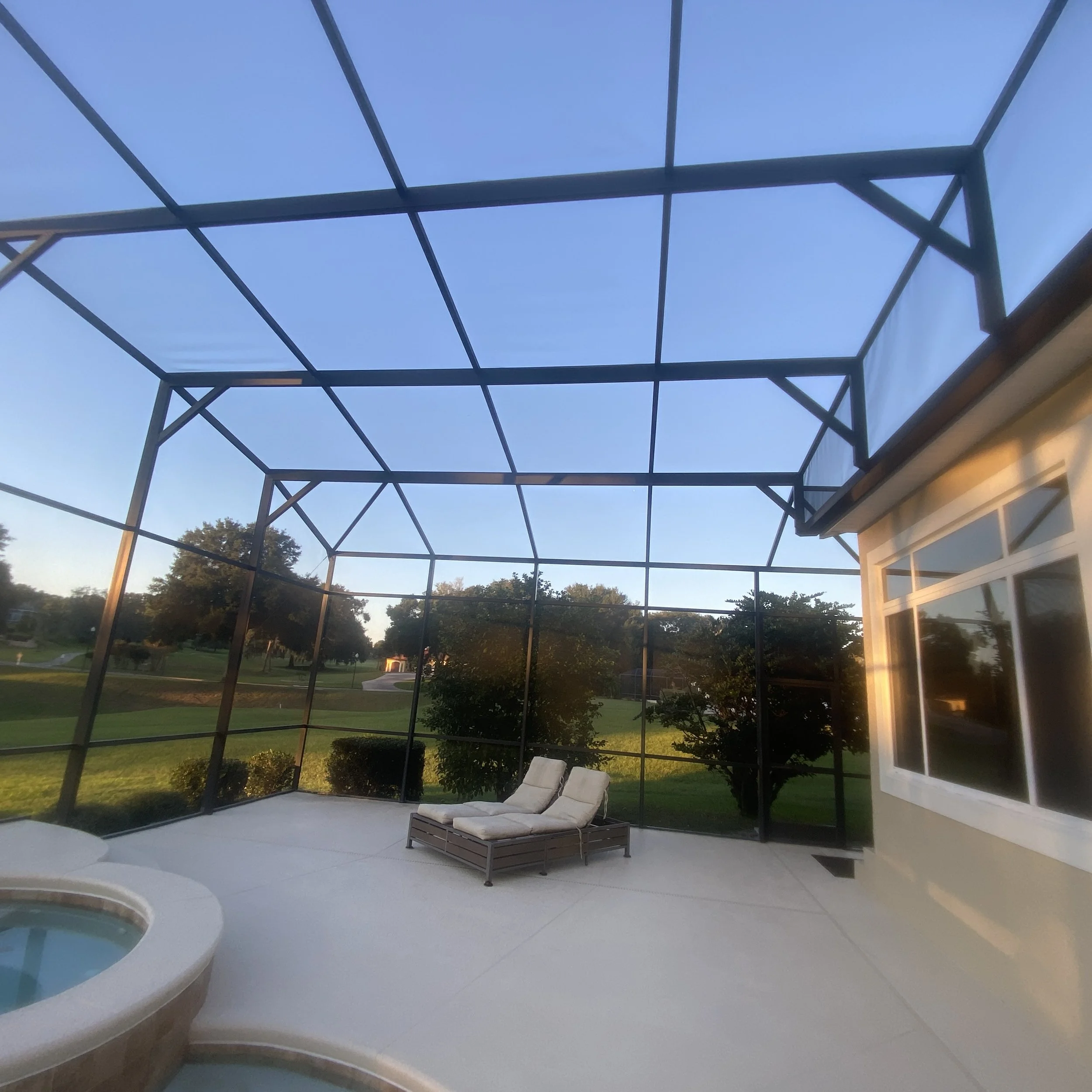 An enclosed backyard patio with a hot tub, two lounge chairs, and a view of green trees and a lawn outside.