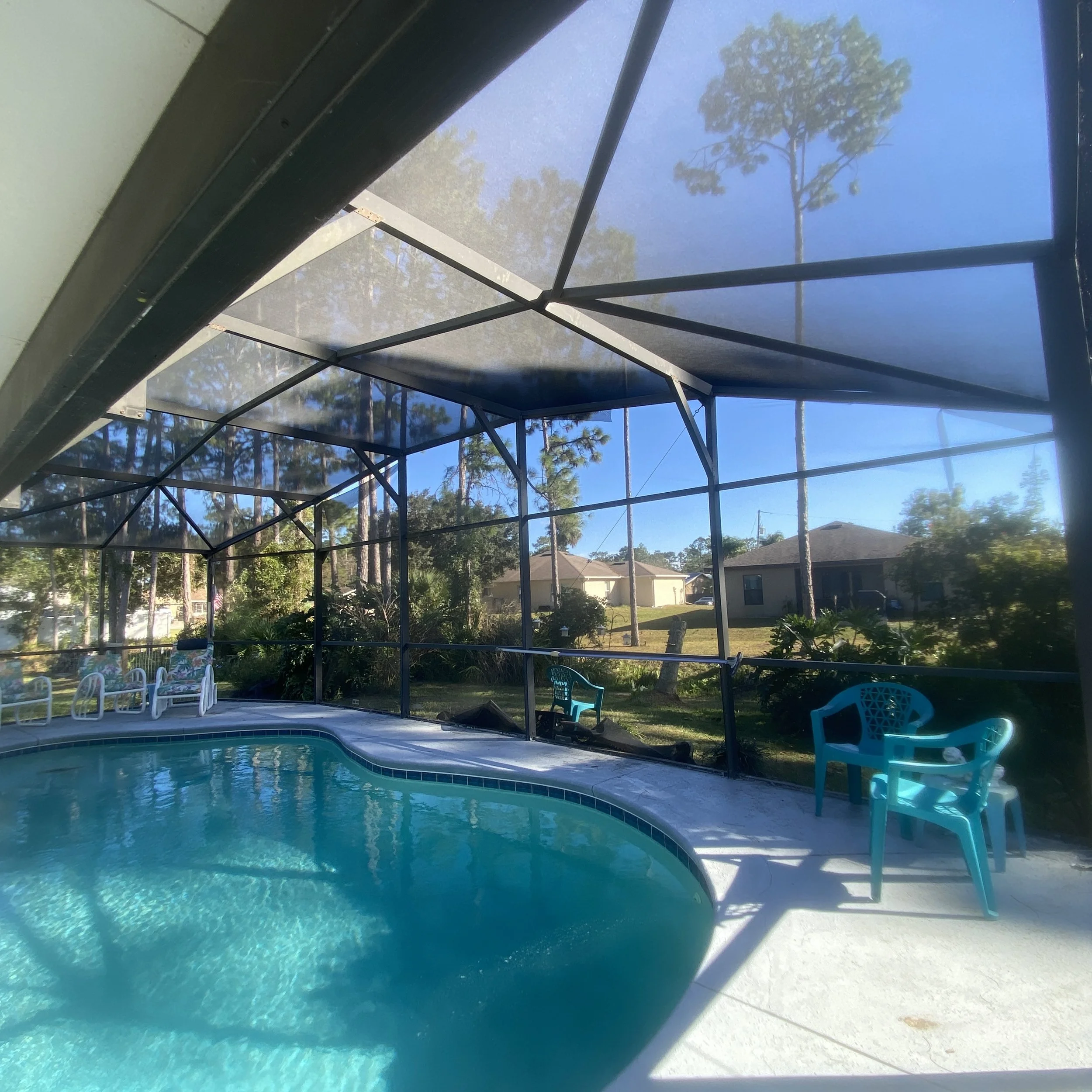 An enclosed backyard swimming pool area with a screened roof, blue water, and plastic chairs in the sun.