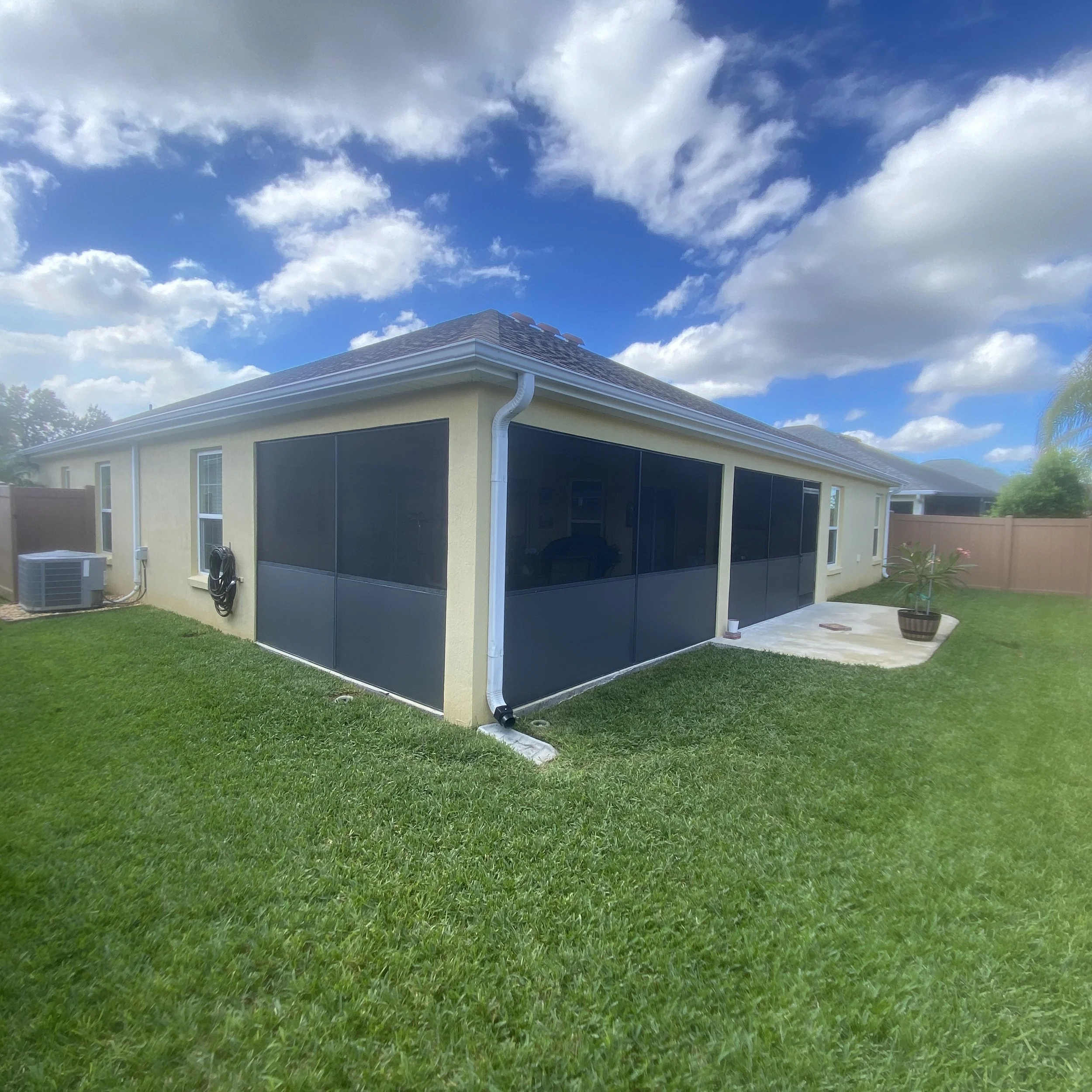 Backyard of a house with a screened porch, green grass, potted plant, and an air conditioning unit, under partly cloudy sky.