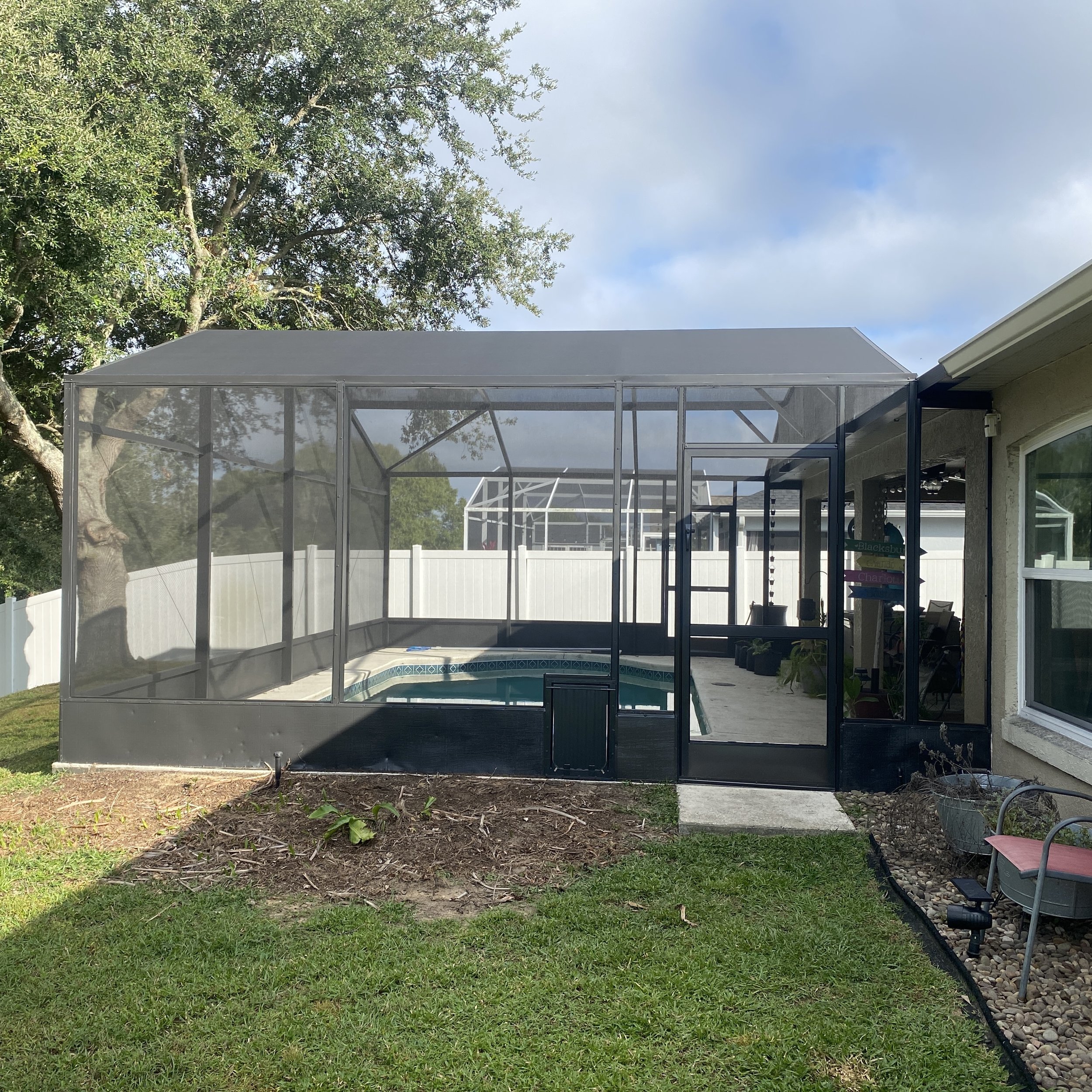 Backyard with screened patio enclosure featuring a swimming pool, a concrete pathway, a garden bed with mulch and plants, and a white fence surrounding the area.