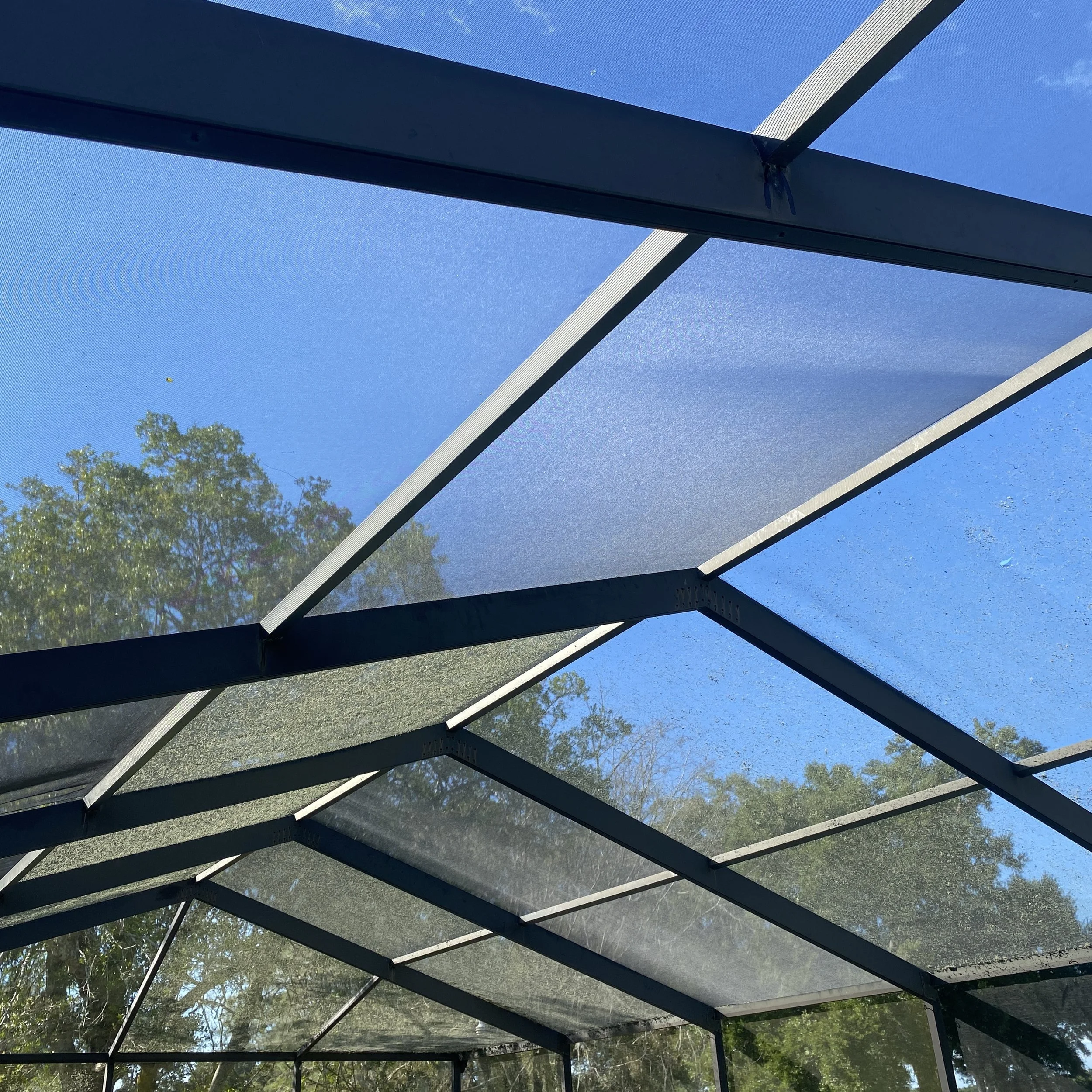 View from beneath a screened and shaded patio or porch enclosure with metal framing, showing the sky and treetops outside.