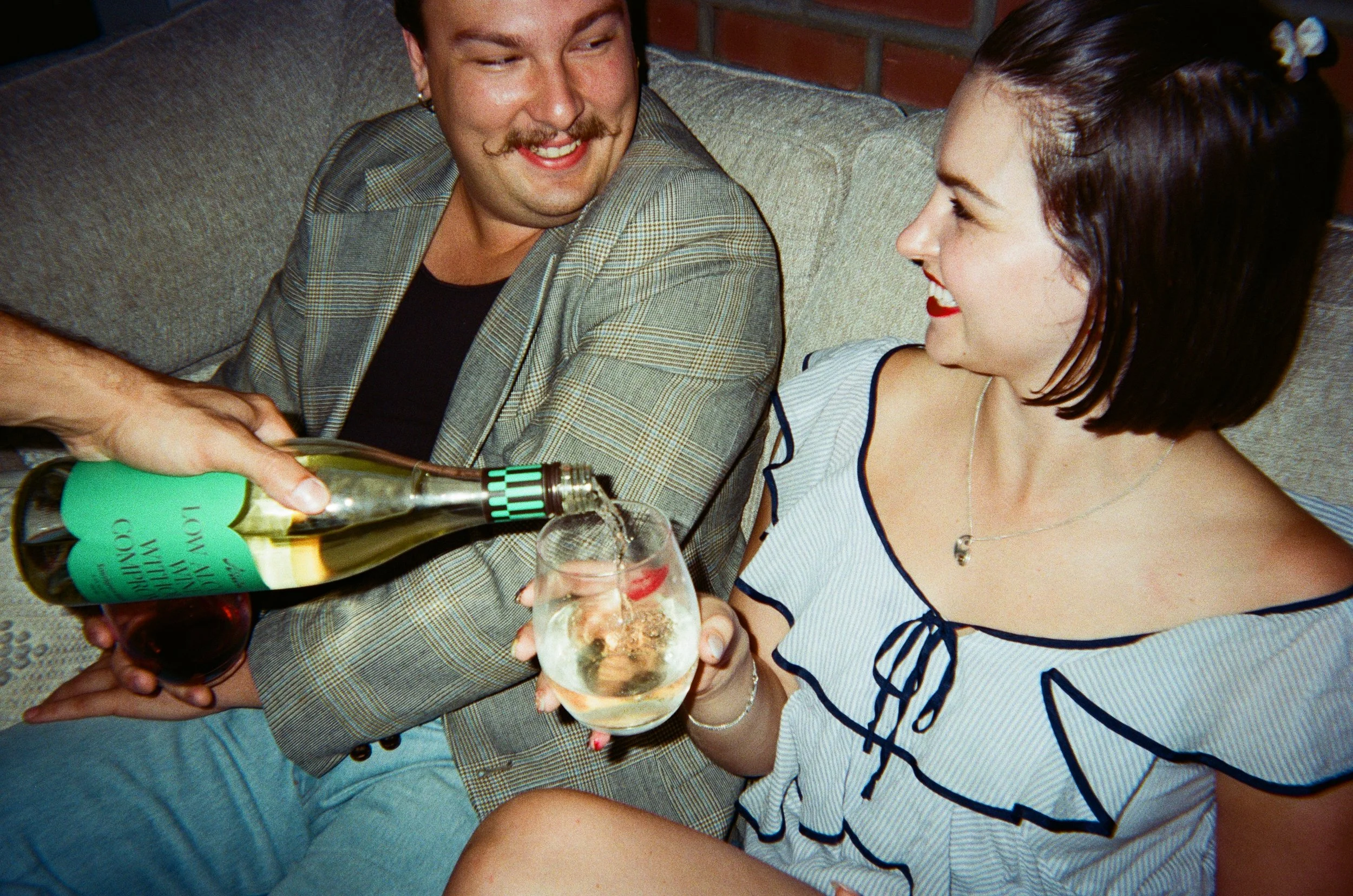 A man and woman smiling on a sofa, while a hand pours white wine into a glass held by the woman.