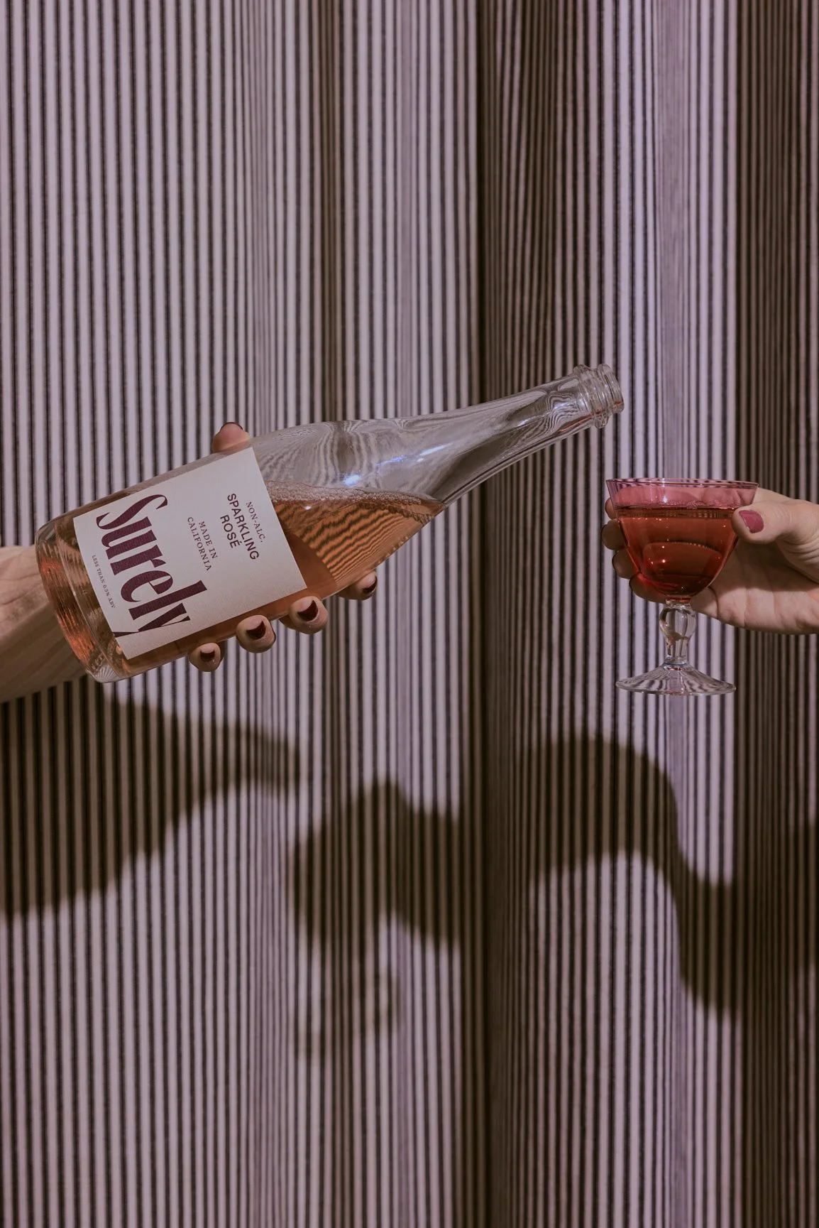 A hand is pouring pink sparkling rosé wine from a bottle into a pink cocktail glass, with a wooden textured background.