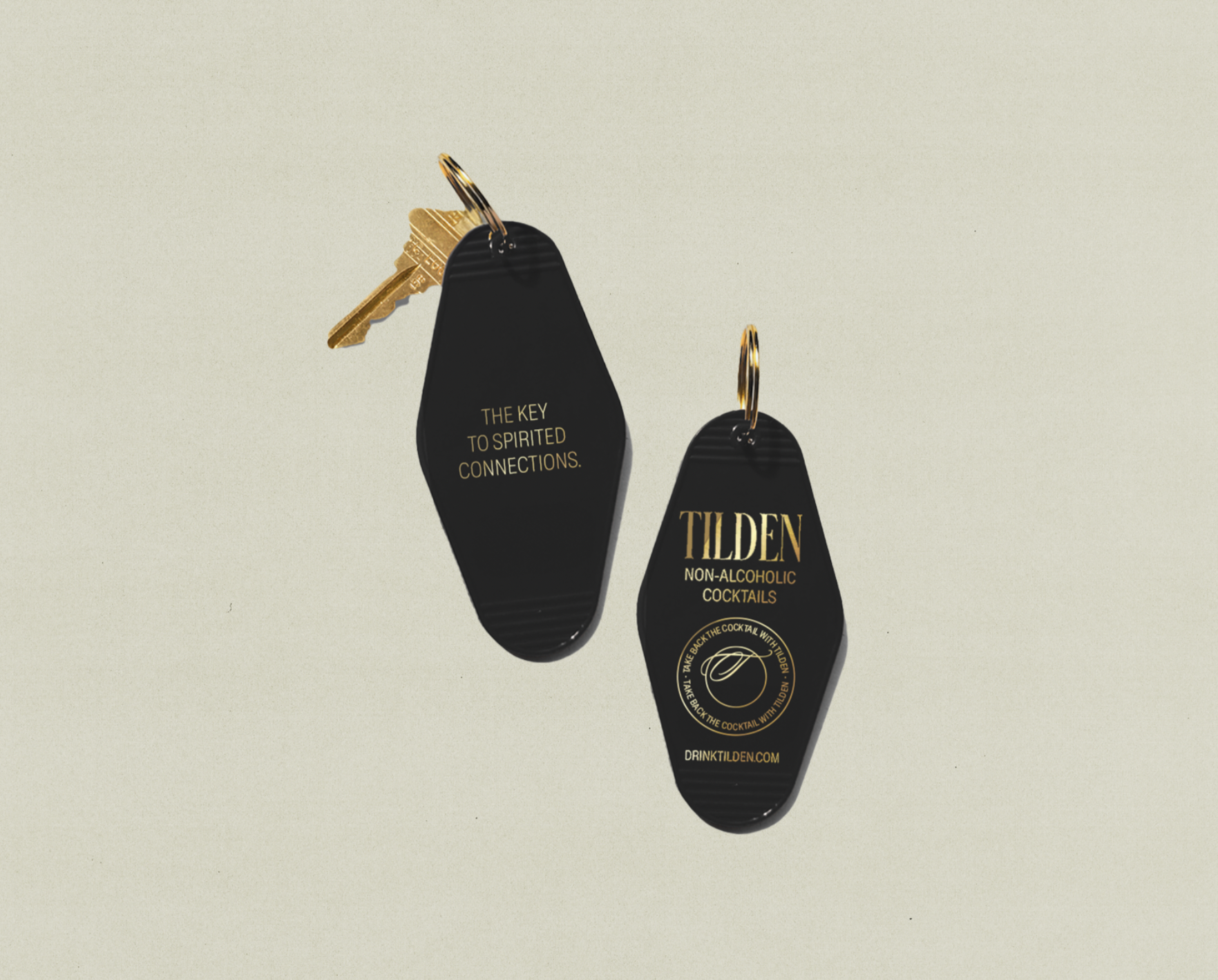 Two black keychains with gold text, one attached to a key, against a plain light background. One keychain says "THE KEY TO SPIRITED CONNECTIONS," the other says "TILDEN NON-ALCOHOLIC COCKTAILS" with a logo and website.