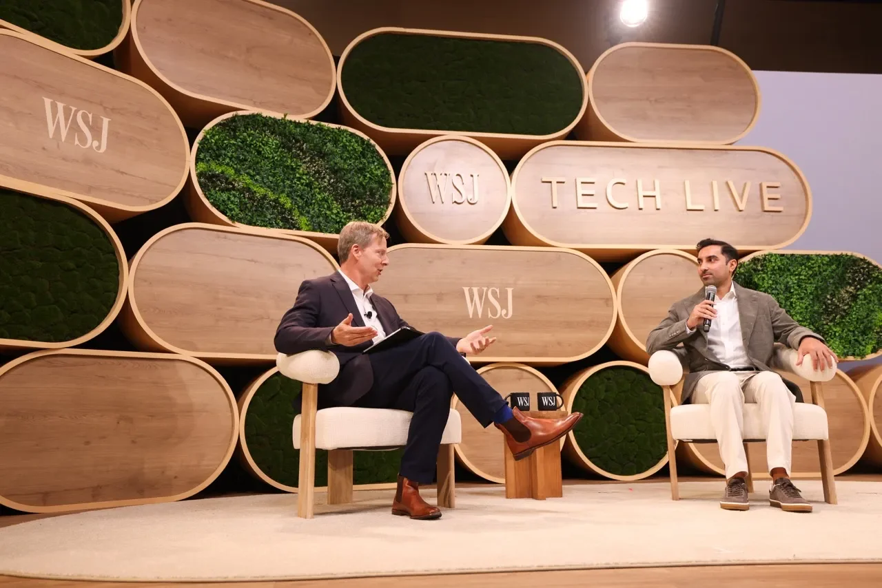 Two men speaking in a panel discussion at WSJ Tech Live event, sitting on stage with modern decorative background of wooden oval shapes and green plants.