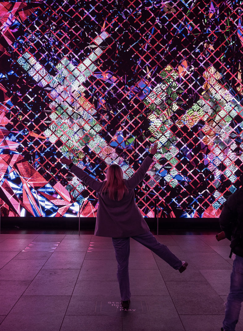 Person standing in front of a large, colorful digital display with arms outstretched and one leg raised in an indoor exhibit.