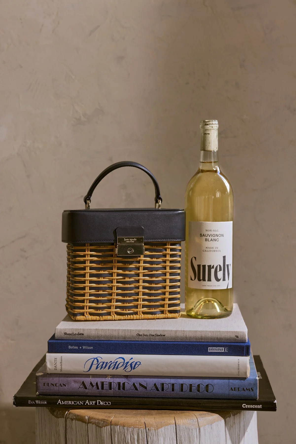 A stack of five art and design books on a small wooden stool, with a wicker handbag and a bottle of white wine on top of the books. The books are titled "American Art Deco," "Paradise," "Birkhauser," "One Sun, One Shadow," and "Crescent." The wicker handbag has a black leather top handle and clasp, and the wine bottle is labeled "Surely" Sauvignon Blanc.