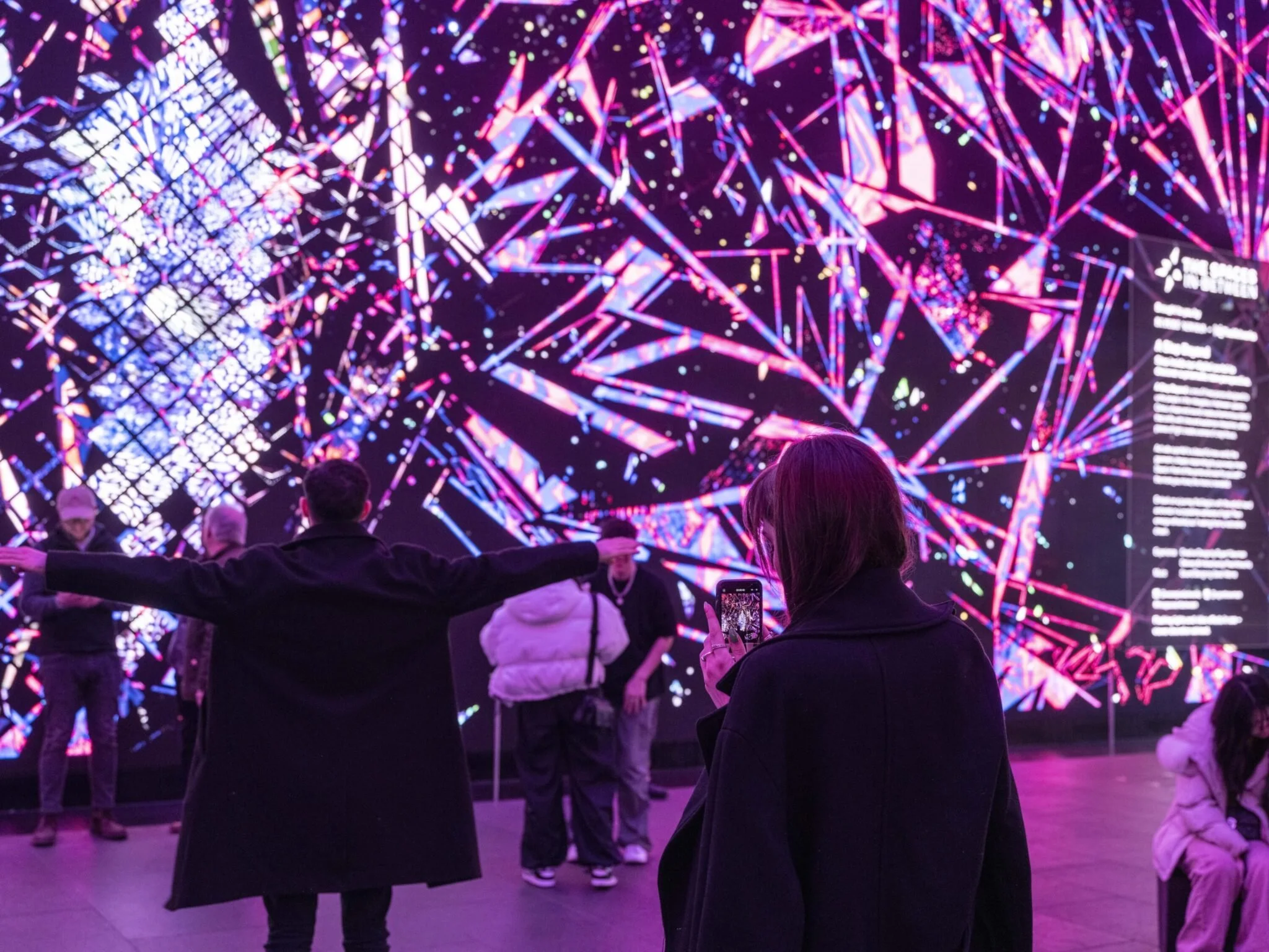 People viewing a vibrant digital art installation with pink, purple, and blue geometric patterns and text on the right.