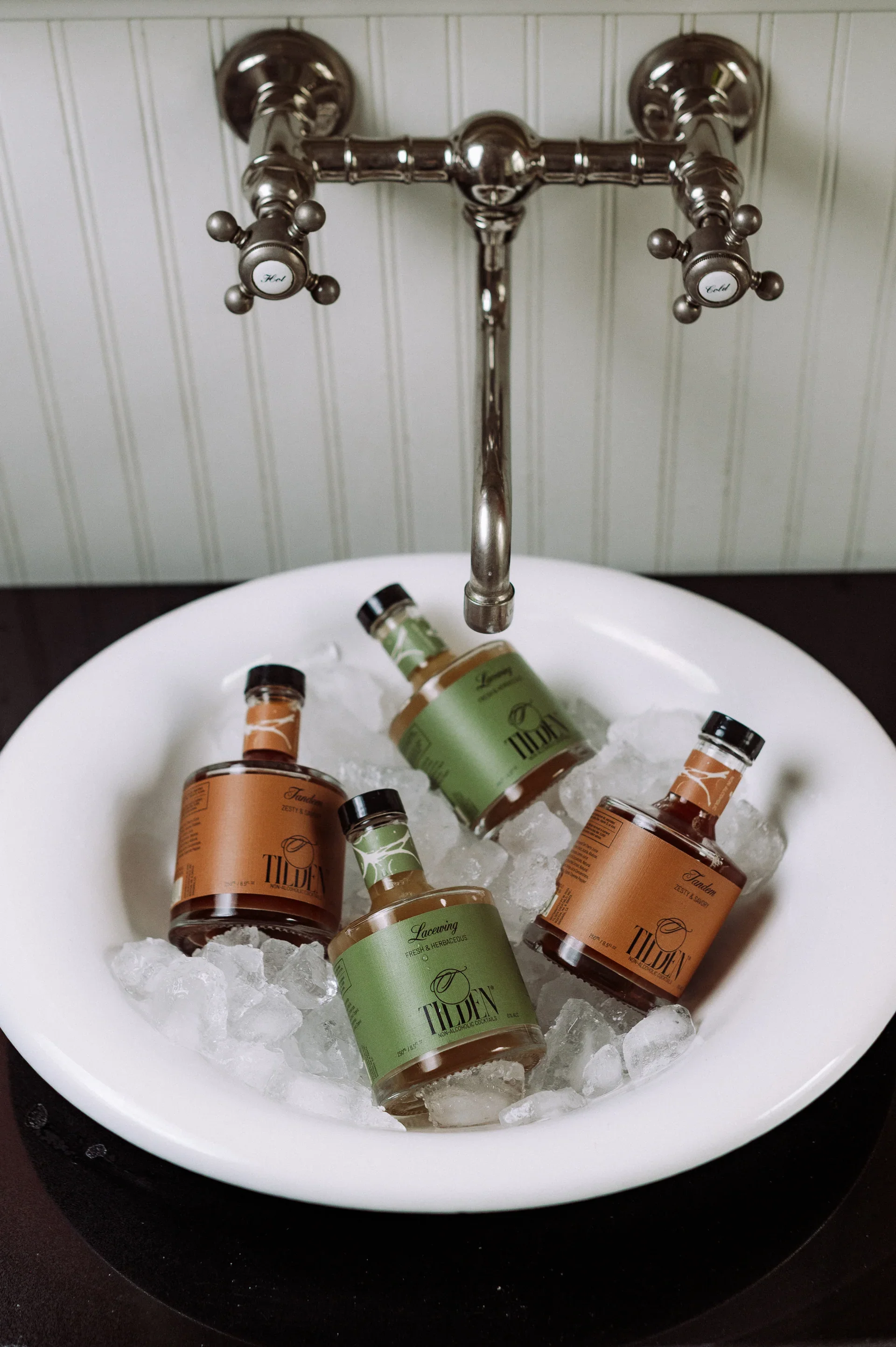 Six jars of Tilden skincare products with green and brown labels are placed on ice in a white bowl under a vintage silver faucet.