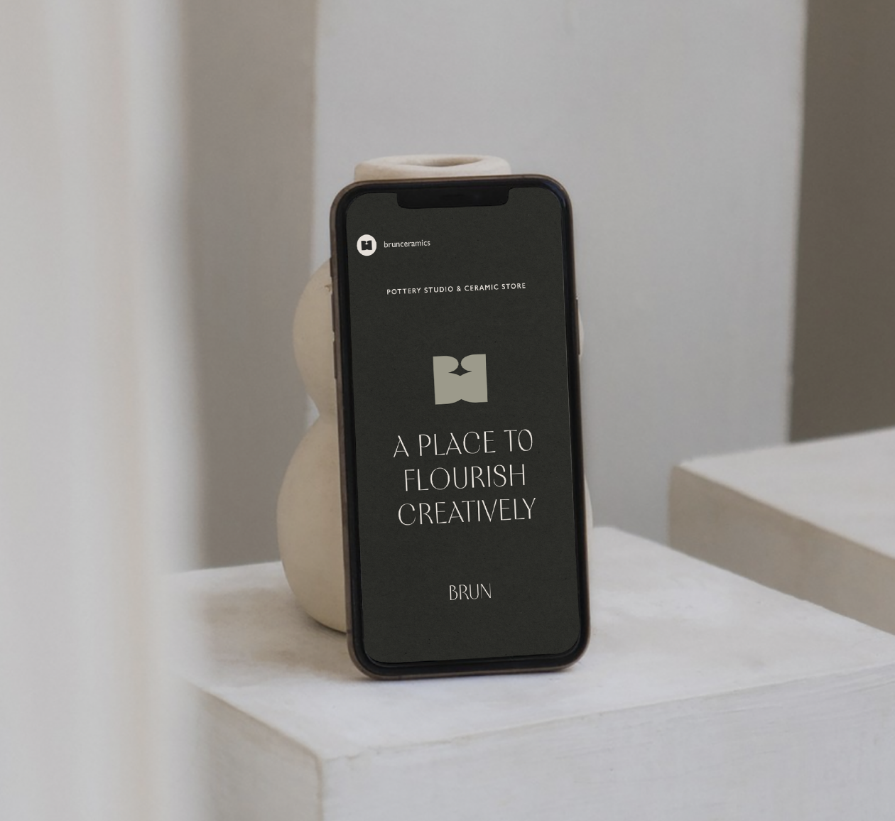 A smartphone displaying a pottery studio and ceramic store's website, placed on a white surface behind a beige, rounded ceramic sculpture. The website's tagline reads, 'A Place to Flourish Creatively.'