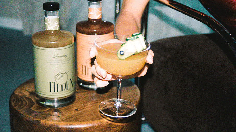 A hand holding a cocktail glass with a peach-colored drink and a cucumber garnish, placed on a wooden table with two bottles of liquor in the background.