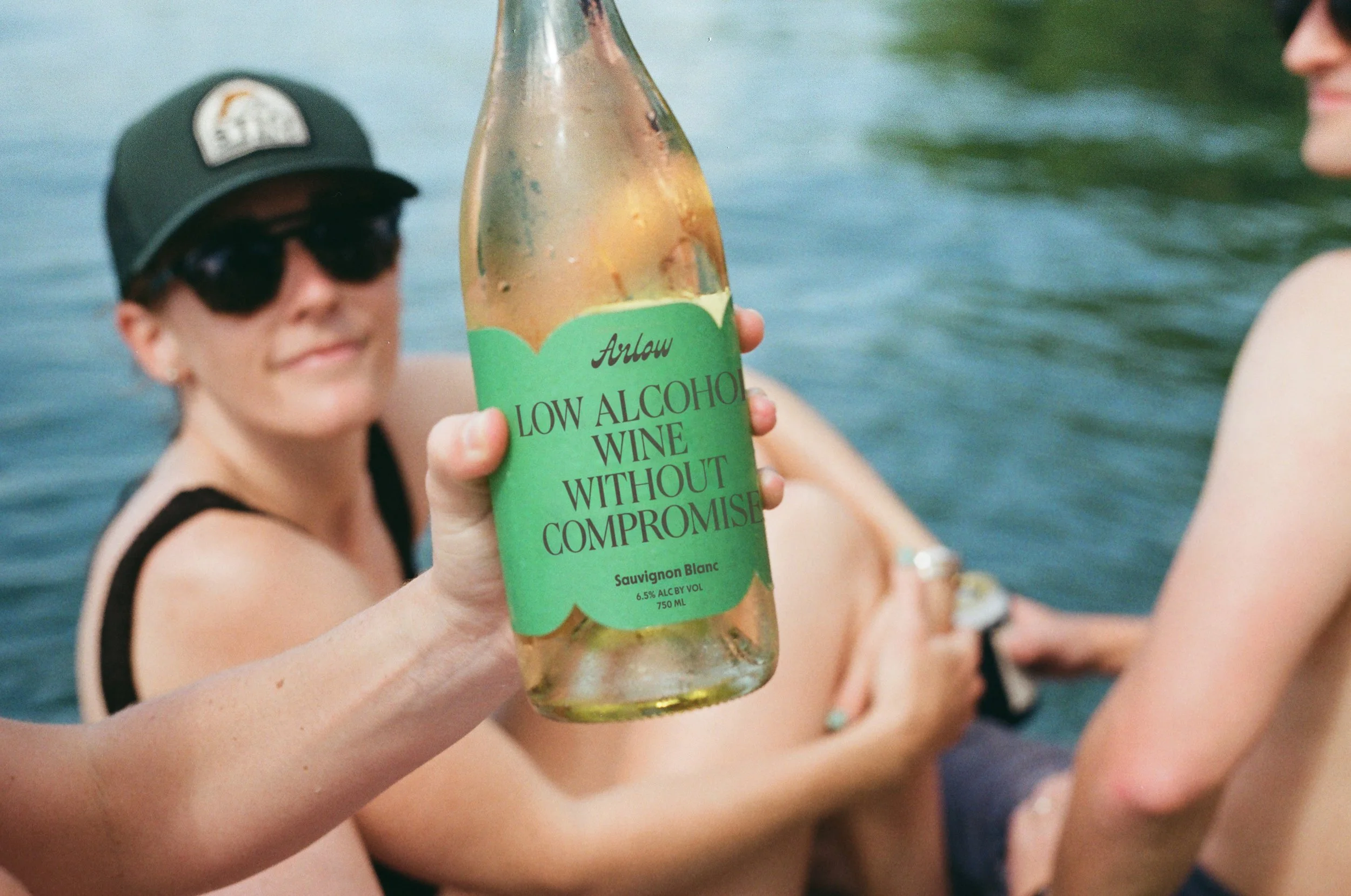 Person holding a bottle of low alcohol wine while sitting in a lake with two other people in the background.