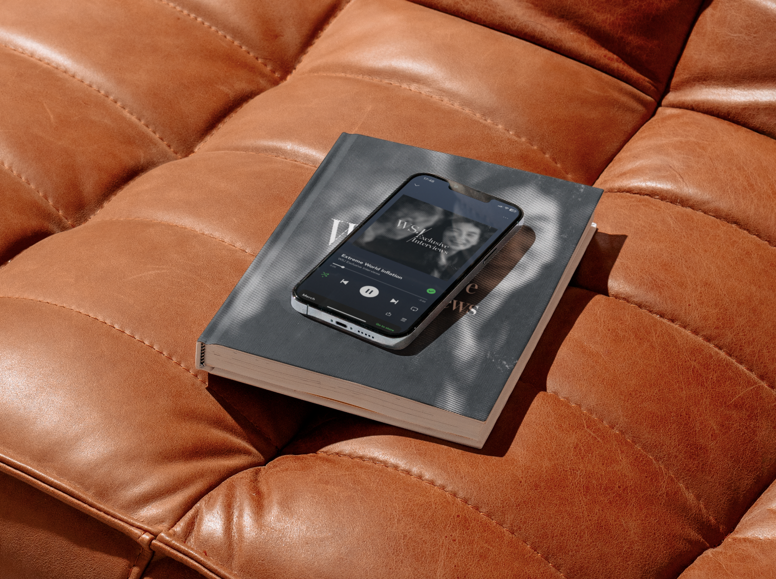 A smartphone playing Spotify music app rests on top of a hardcover magazine or book on an orange leather couch.