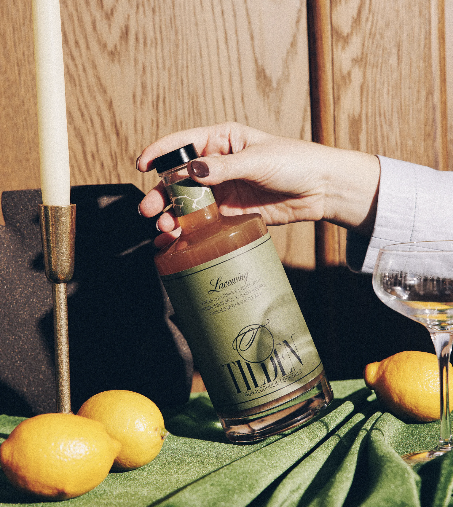 A hand with manicured nails holding a bottle of Lacrew nonalcoholic cocktail, with lemons, a glass of drink, and a candle on a green surface with a wooden background.