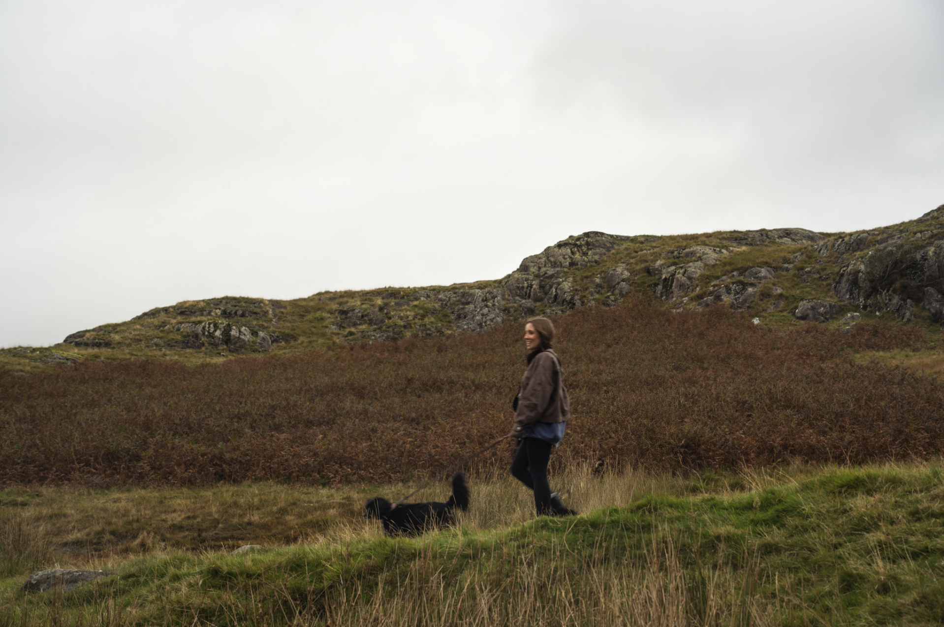 A woman walking her black dog in a grassy, hilly landscape with a rocky hill in the background under an overcast sky.