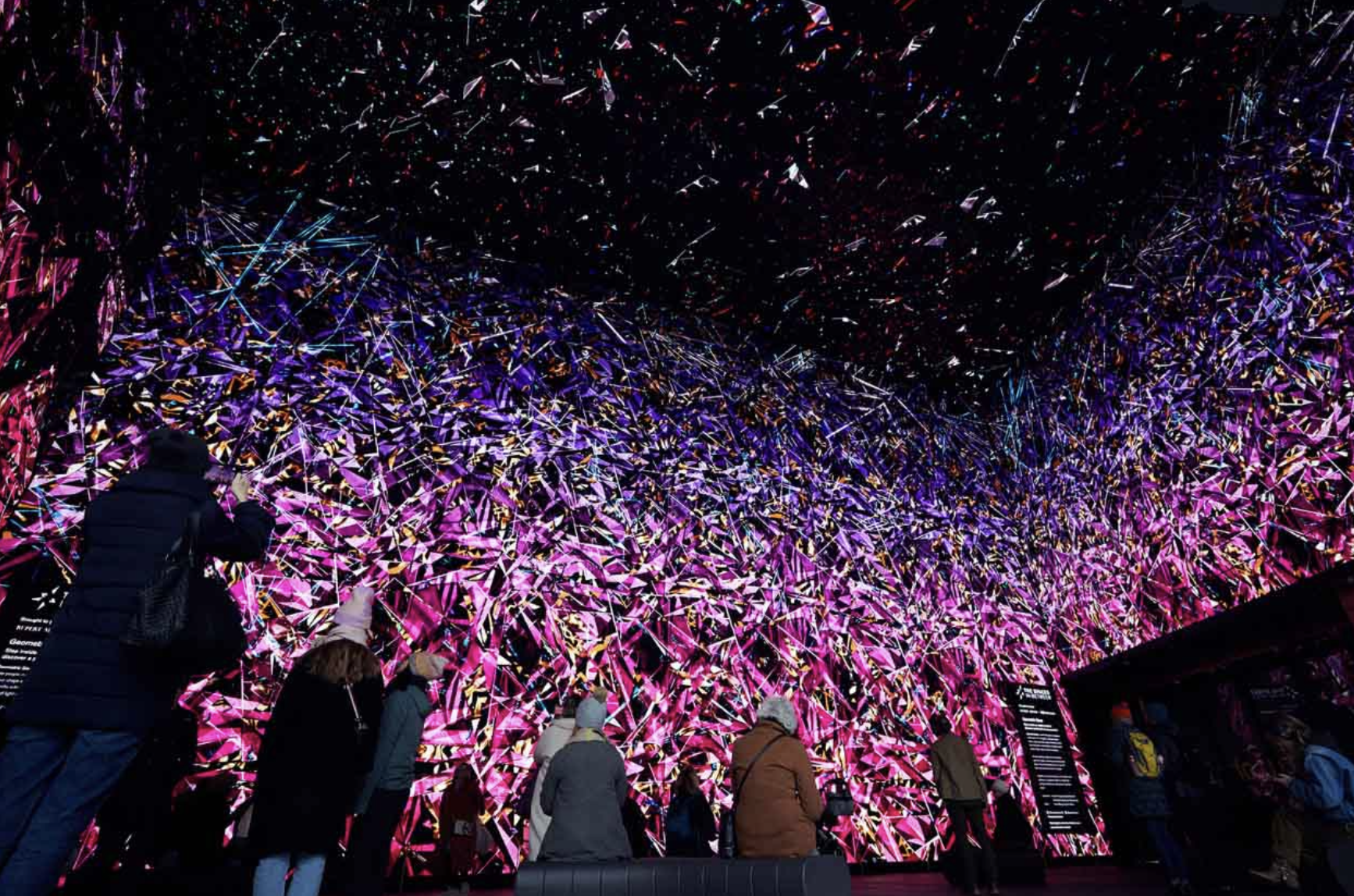 People observing a large digital art installation featuring a vibrant display of colorful geometric patterns and lines on a wide screen.