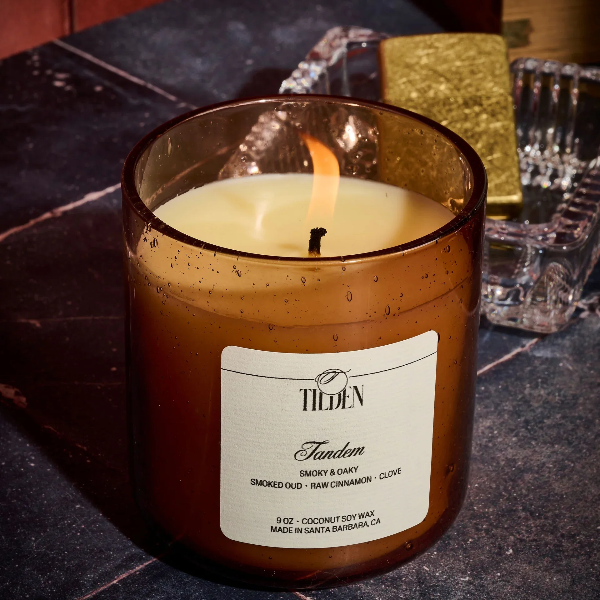 A lit candle in a brown glass container with a white label that reads "Tilden," placed on a dark surface, with a gold and clear decorative object and a clear glass holder in the background.