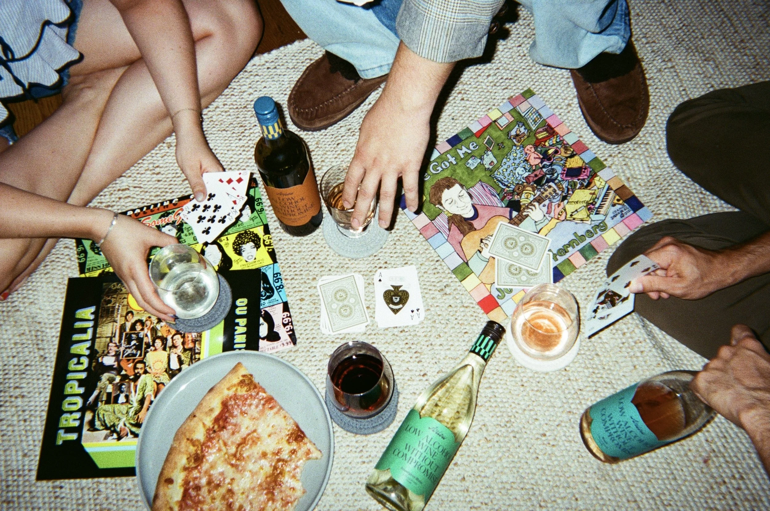 Group of people playing cards, drinking wine and beer, with a pizza on a carpeted floor.