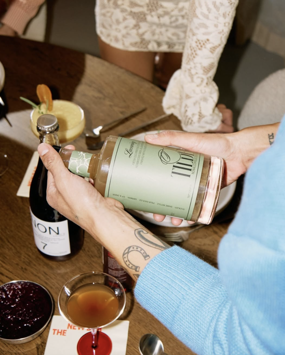 Person opening a bottle of rosé wine at a table with drinks and desserts, including a glass of amber-colored drink and bowls of berry compote.