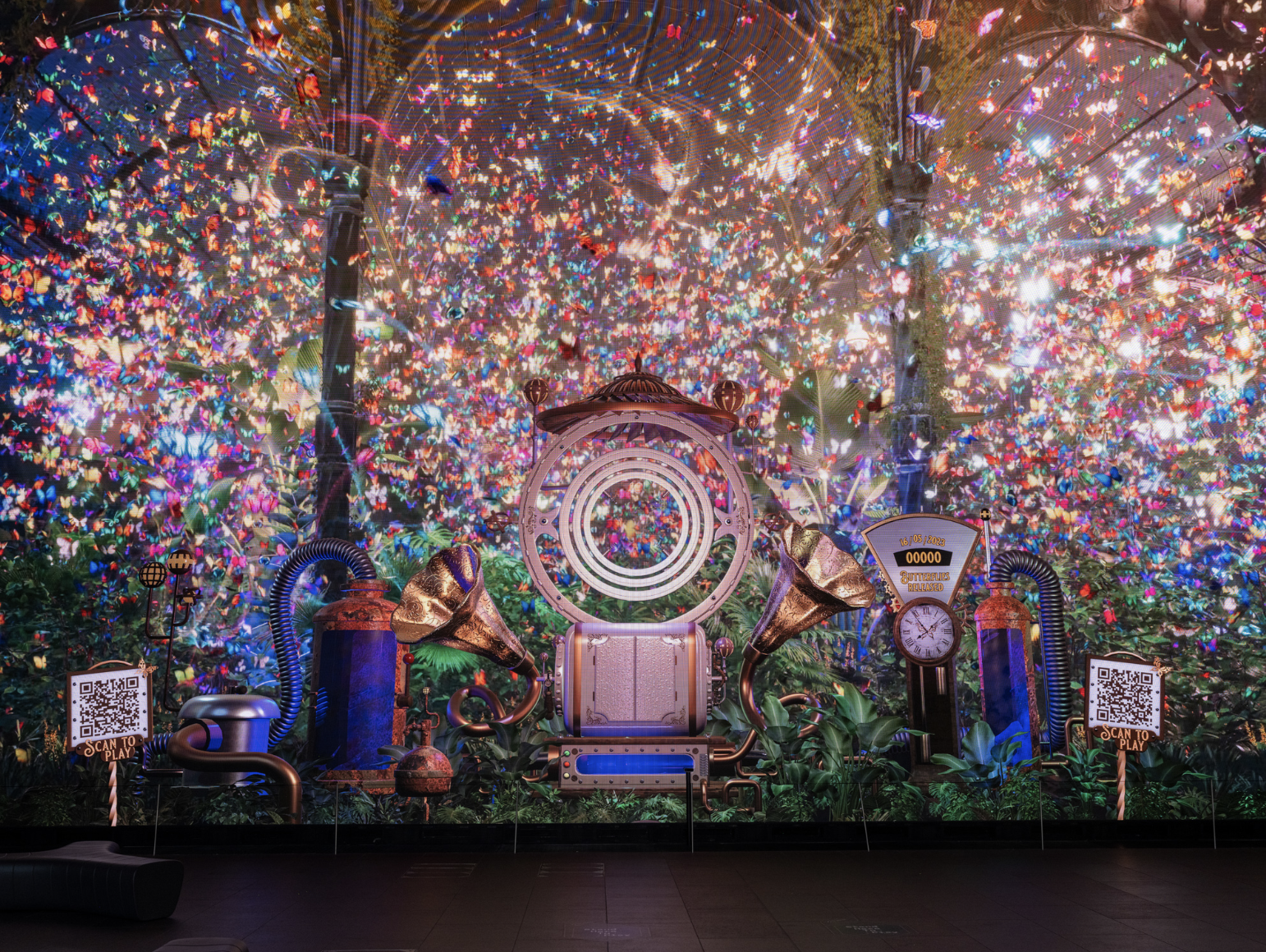 An art installation resembling a whimsical, metallic machine with large horns, surrounded by colorful butterflies, plants, and QR codes on signage, set against a vibrant backdrop of shimmering, multicolored lights and butterflies.