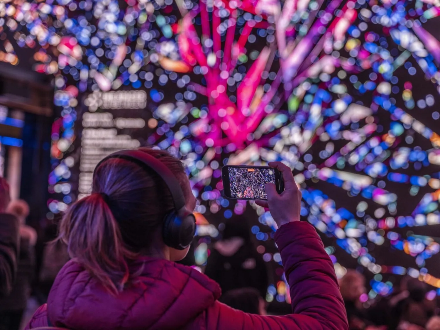 A woman with a ponytail wearing headphones and a red jacket is taking a photo with her phone of a brightly lit, colorful tree made of lights.