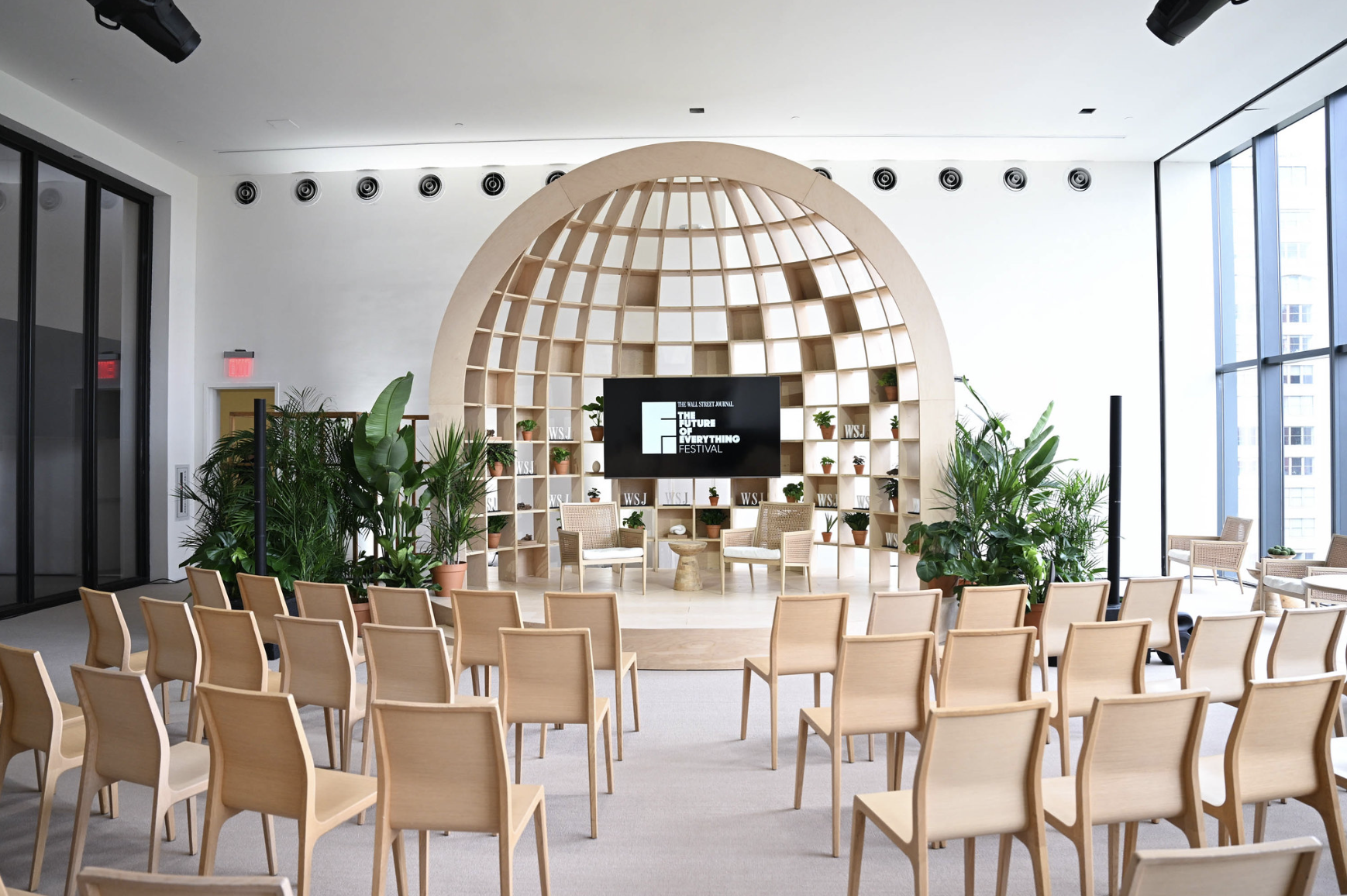 Modern conference room with a semi-circular wooden stage, chairs arranged in rows, potted plants, large windows, and a digital screen displaying 'The Future of Everything Festival'