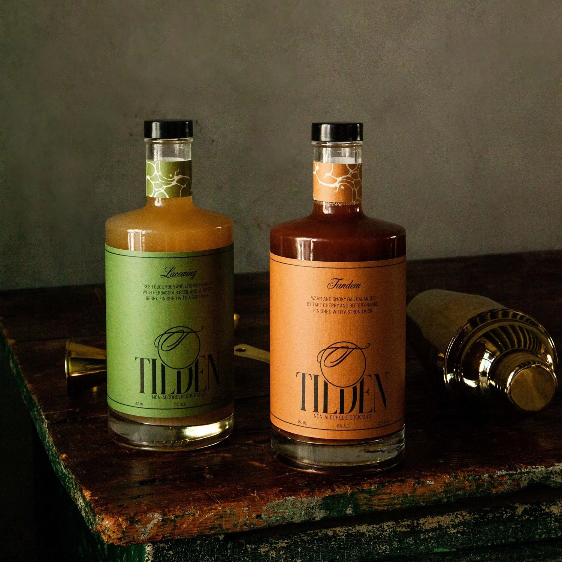 Two bottles labeled Tilden non-alcoholic cocktails, one with a green label called 'Lacewing' and the other with an orange label called 'Tandem,' placed on a rustic wooden surface with a metallic shaker in the background.