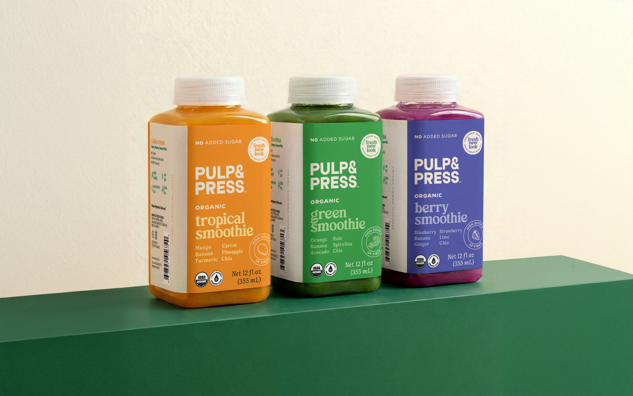 Three bottles of Pulp & Press organic smoothies in orange, green, and purple bottles, labeled tropical, green, and berry smoothies, on a green surface.