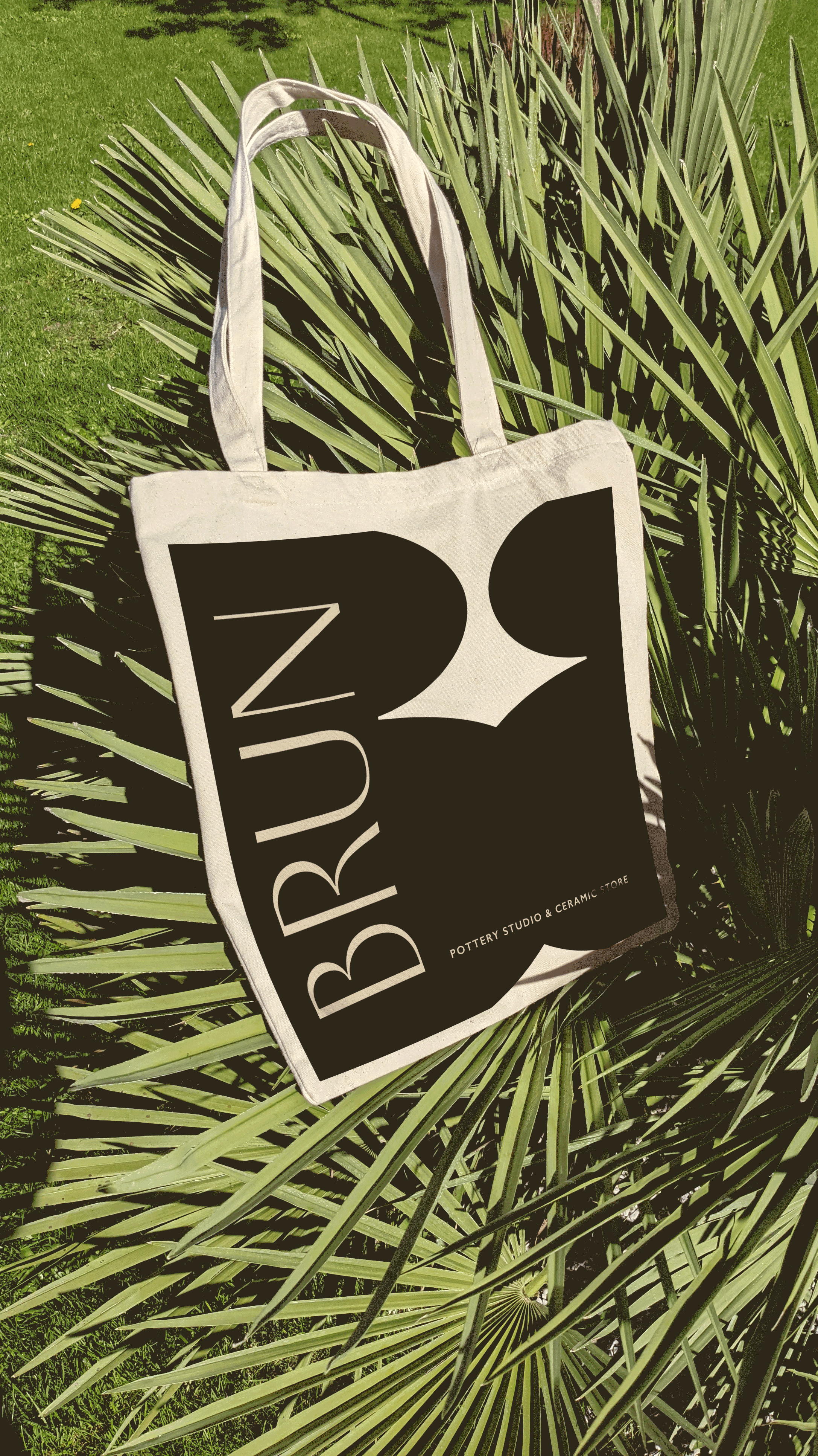 A beige tote bag with black graphic resembling the word 'BROWN' and a stylized bird, resting on green palm leaves outdoors.