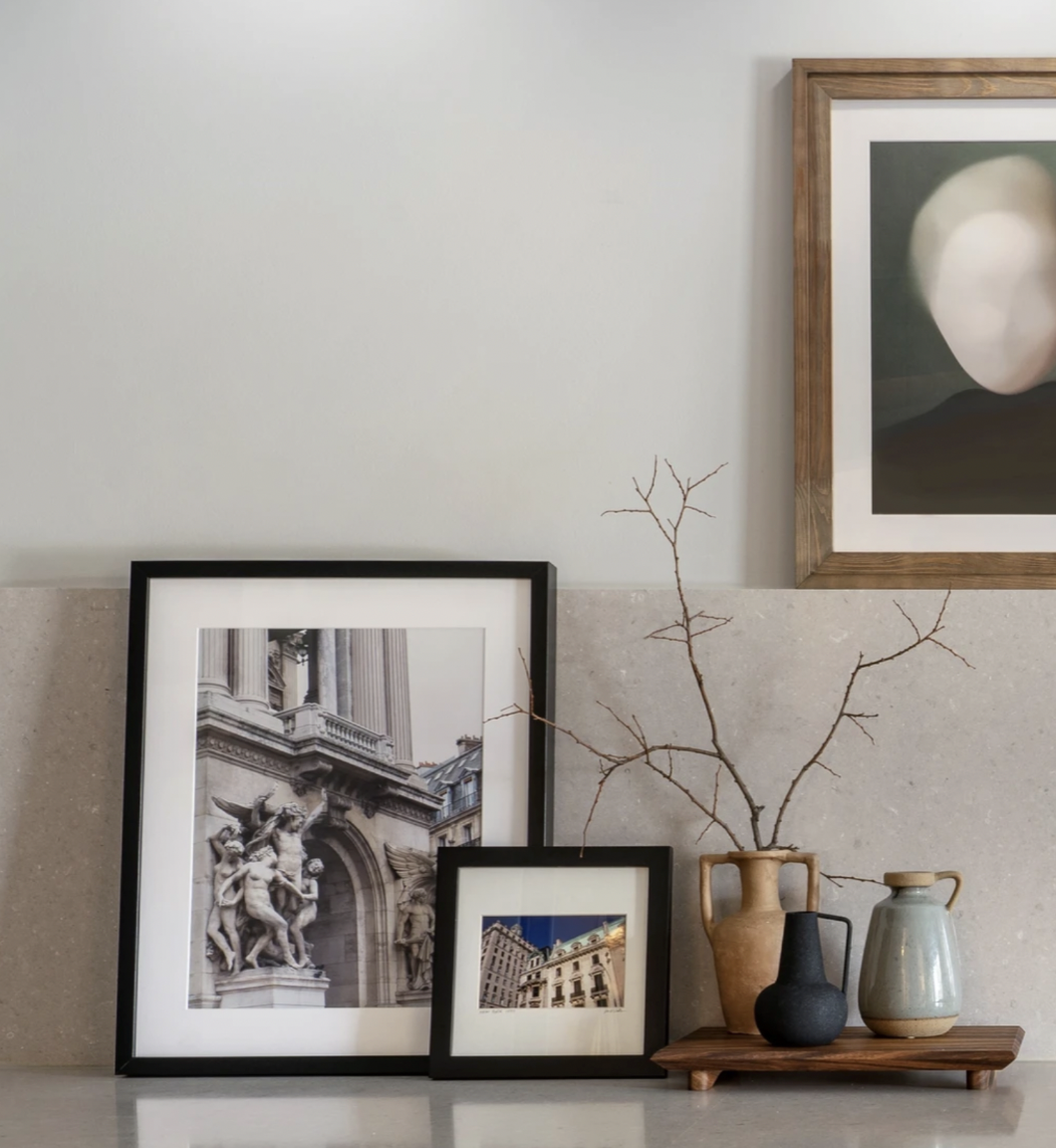 Decorative home interior with framed photographs of classical architecture, a wooden shelf with three ceramic vases and a bare tree branch, and a large framed abstract art piece on the wall.