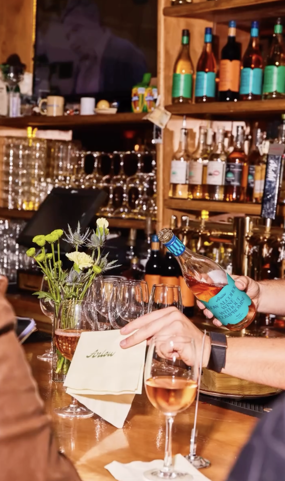 A person is pouring rosé wine into a glass in a bar setting with bottles and glasses on wooden shelves in the background.