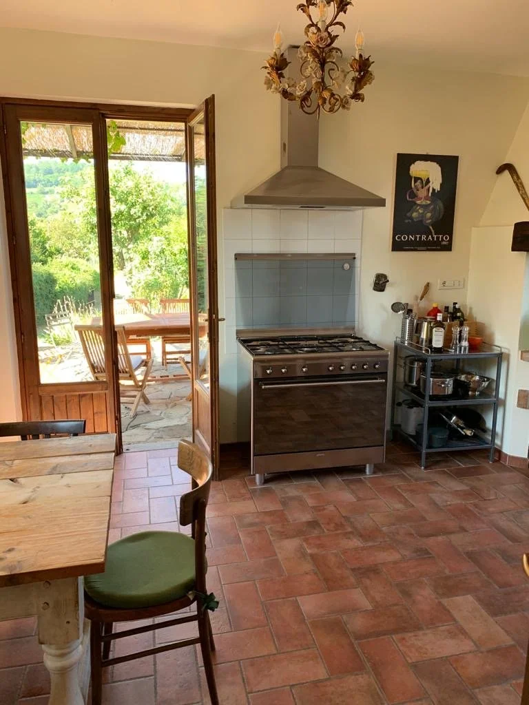 A kitchen with a door leading to an outdoor patio, a stainless steel stove, a black metal cart with bottles and kitchen items, a wooden dining table, and chairs, with terracotta tiled floors and a chandelier hanging from the ceiling.