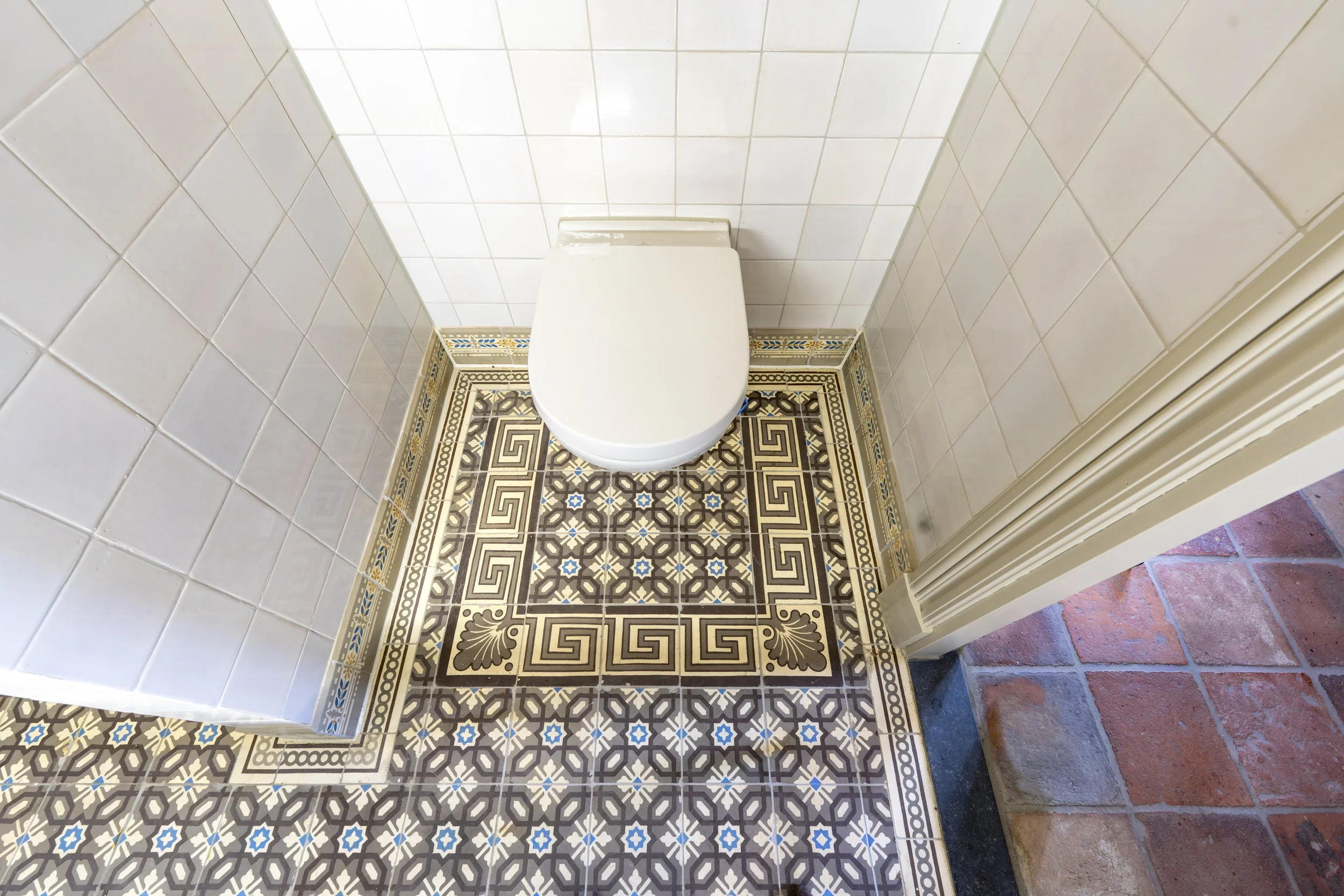 The image shows a toilet in a bathroom with intricate patterned floor tiles and plain white wall tiles.