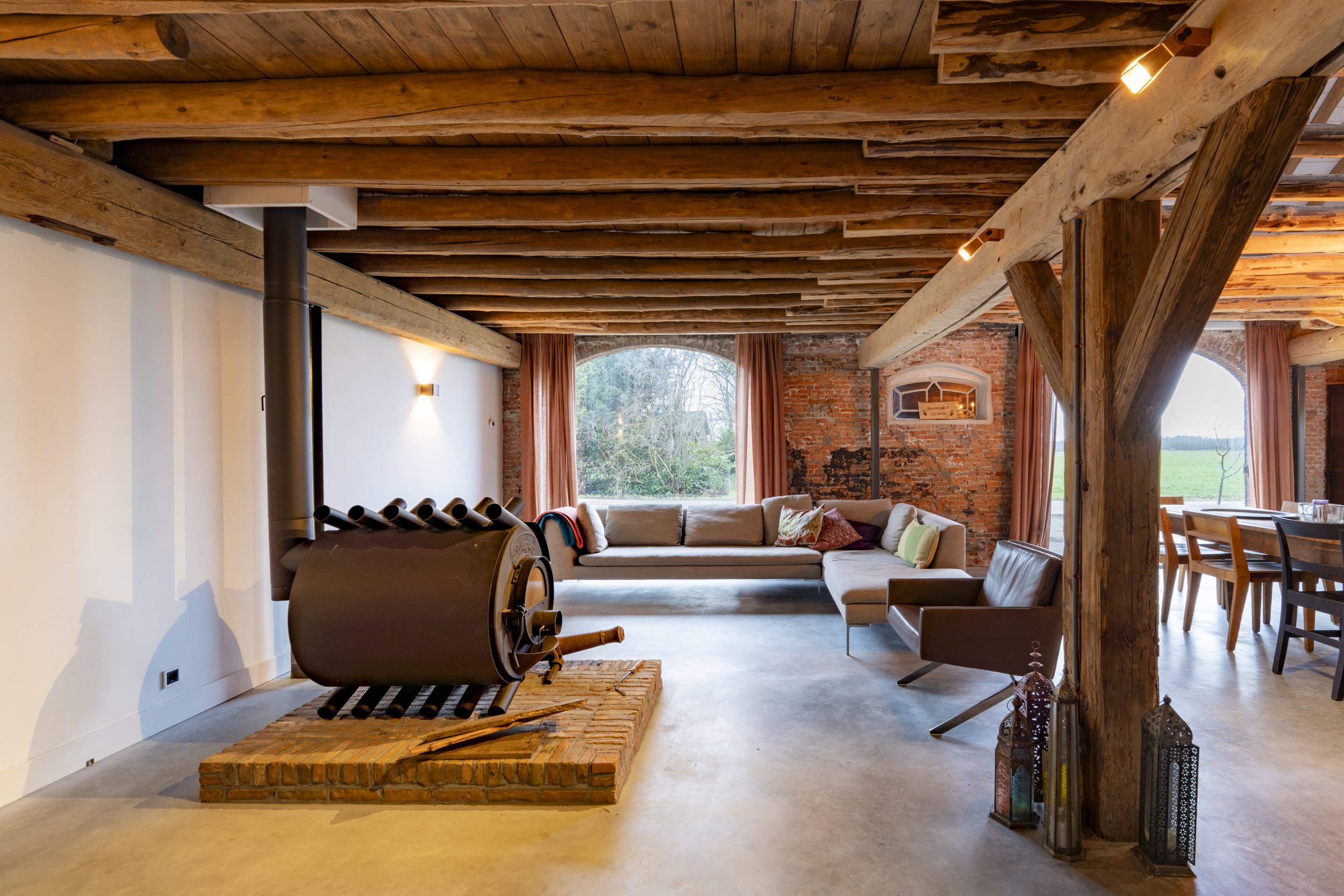 Living room with exposed wooden beams ceiling, large windows with curtains, a sectional sofa, a leather chair, a wood stove on a brick platform, and decorative lanterns.