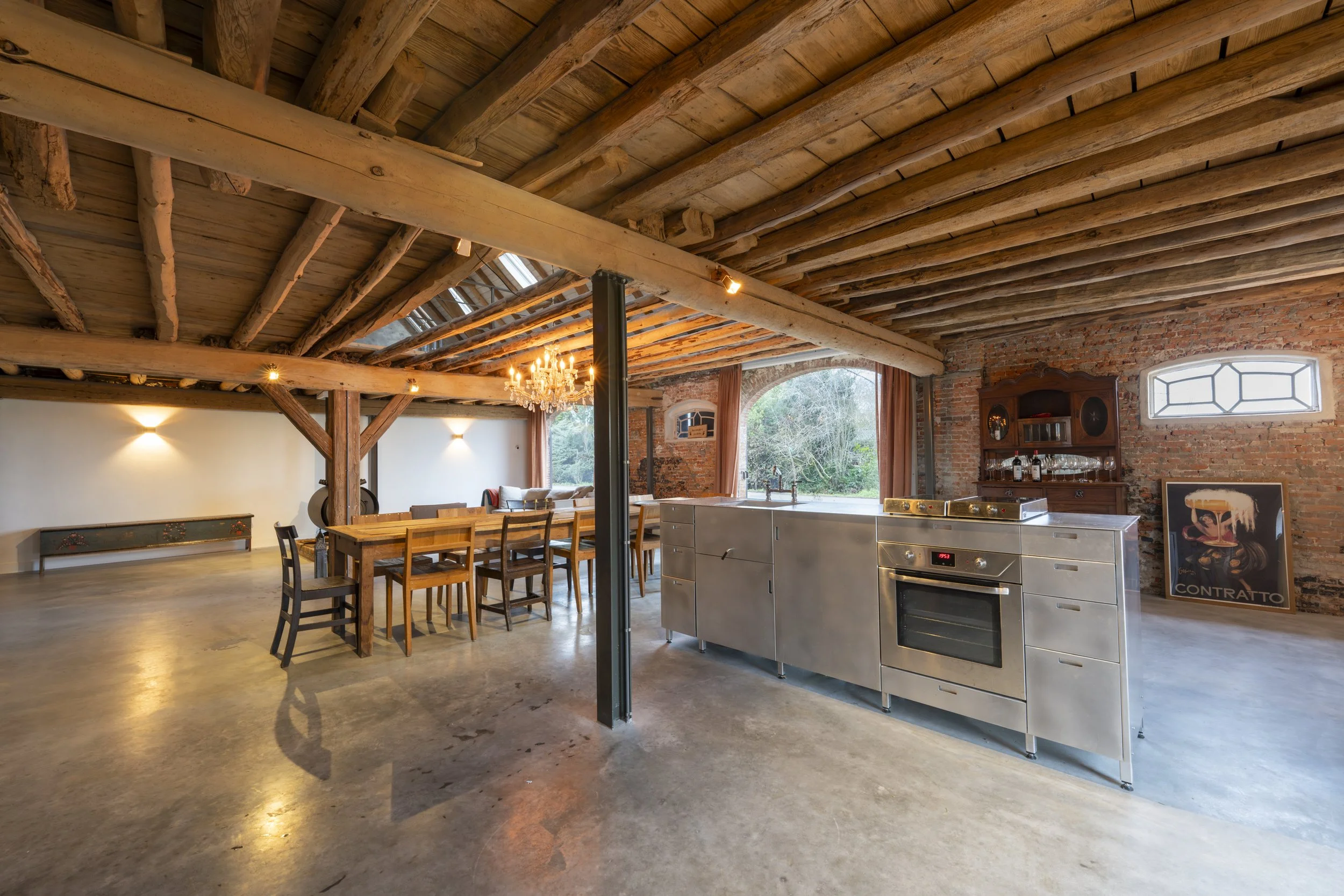 Open-plan rustic kitchen and dining area with exposed wooden beams, a large wooden dining table with chairs, a chandelier, a stainless steel kitchen island, brick walls, and large windows showing greenery outside.