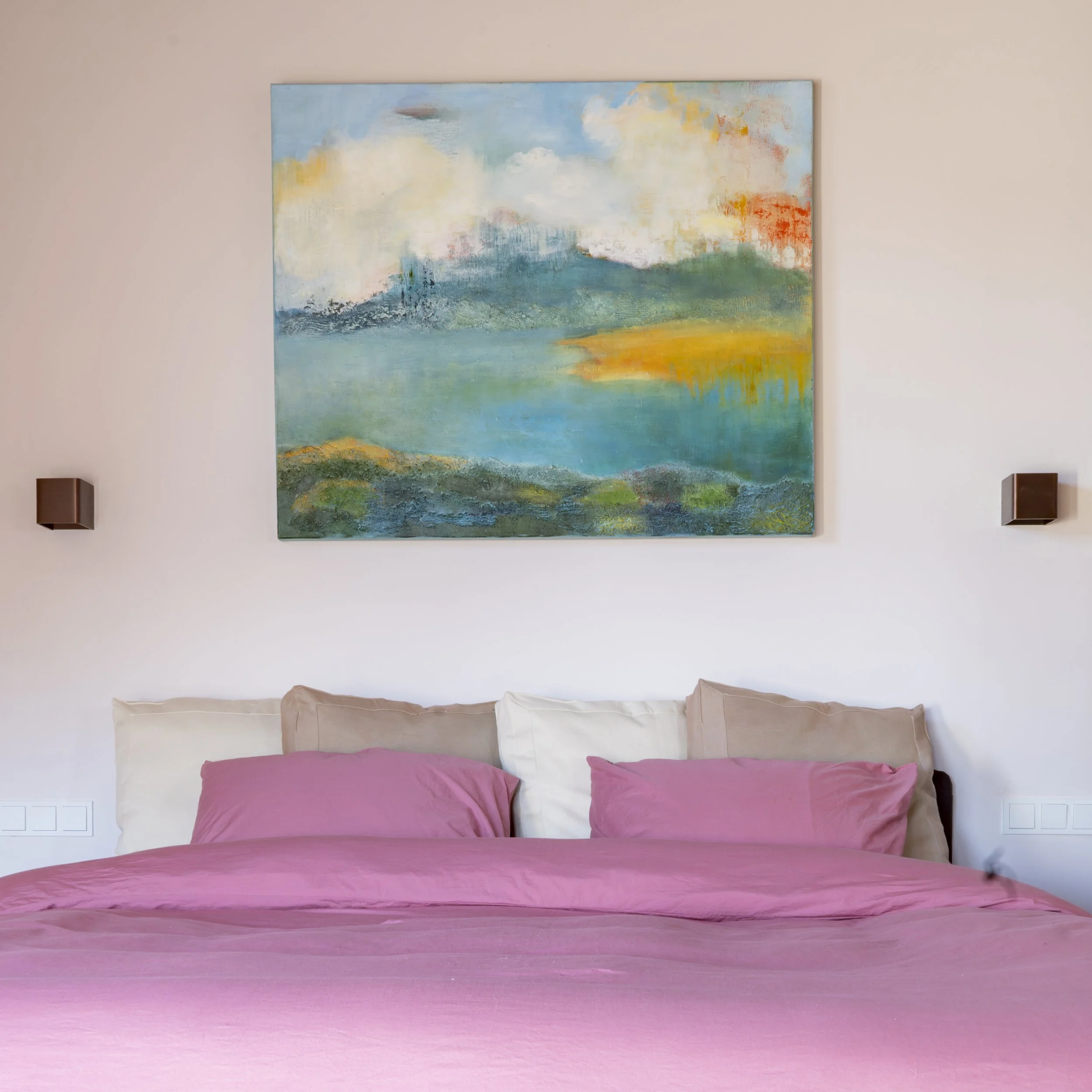 A bedroom with a pink bedspread, multiple pillows in beige to white, a landscape painting above the bed featuring water, greenery, and sky, and wall-mounted light fixtures on each side of the painting.