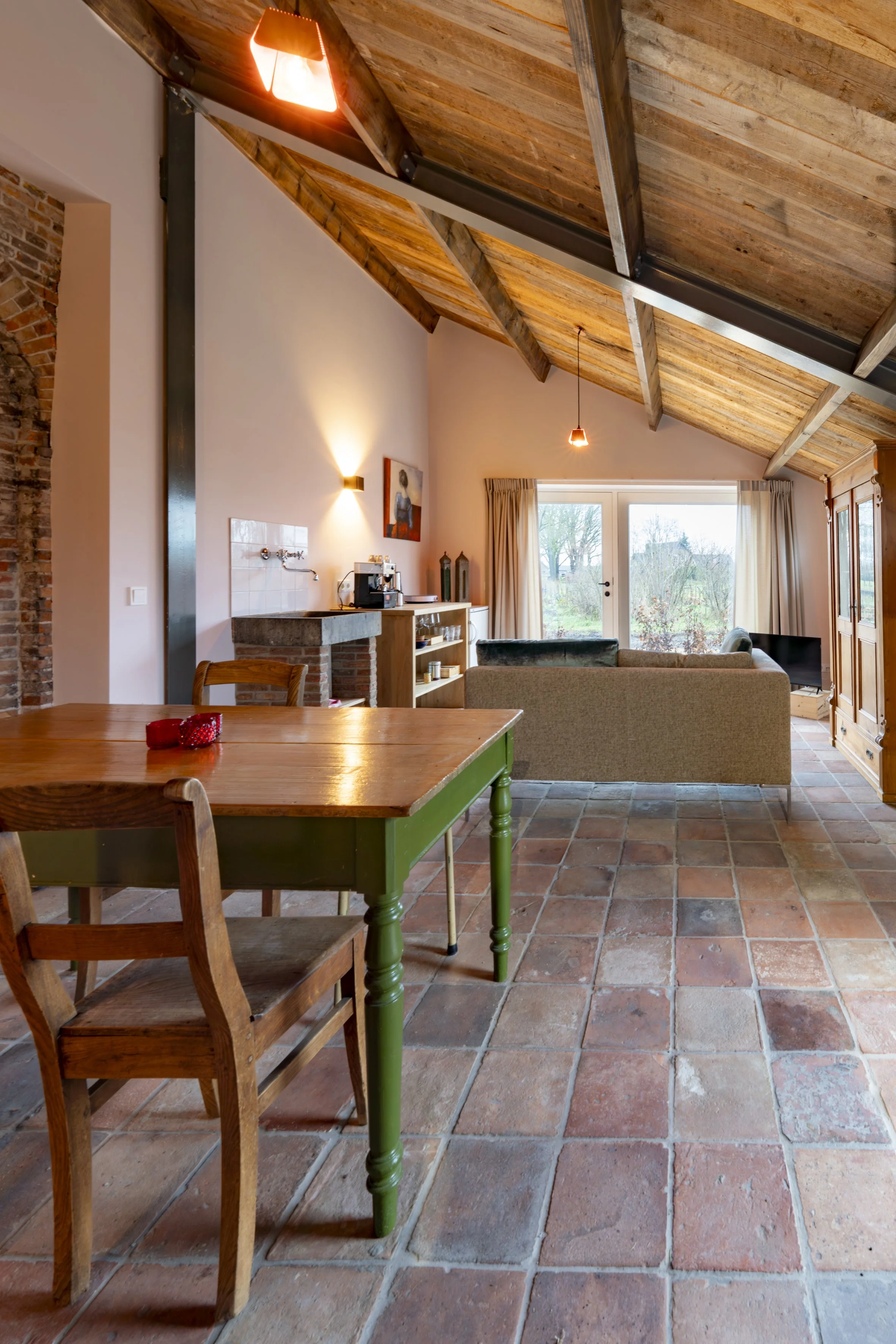 Living room with wooden ceiling, brick wall, tile flooring, dining table, chairs, couch, cabinet, and large window.