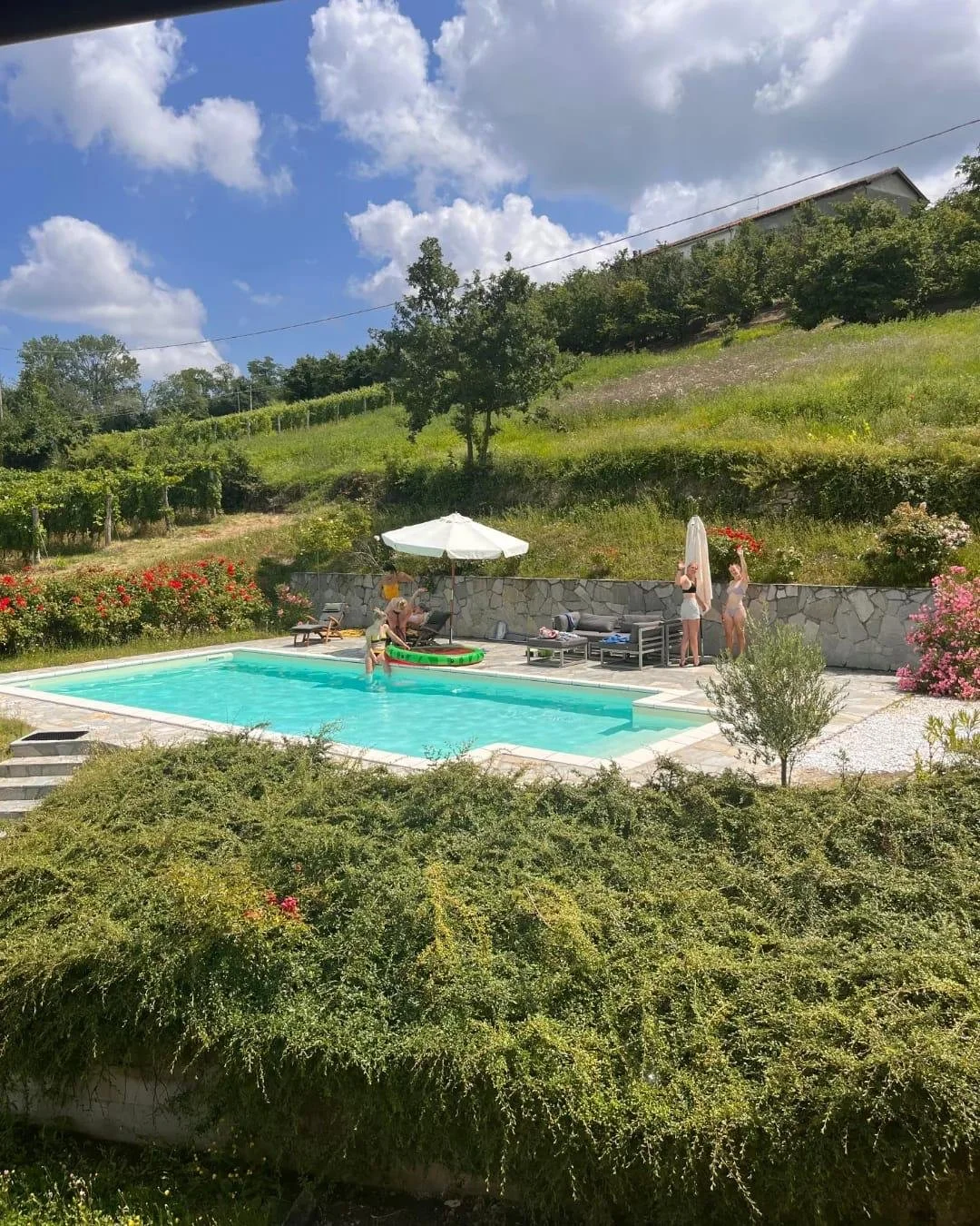 People enjoying a sunny day around a backyard swimming pool with lounge chairs, an umbrella, and a child in the pool, surrounded by lush greenery and flowering bushes.
