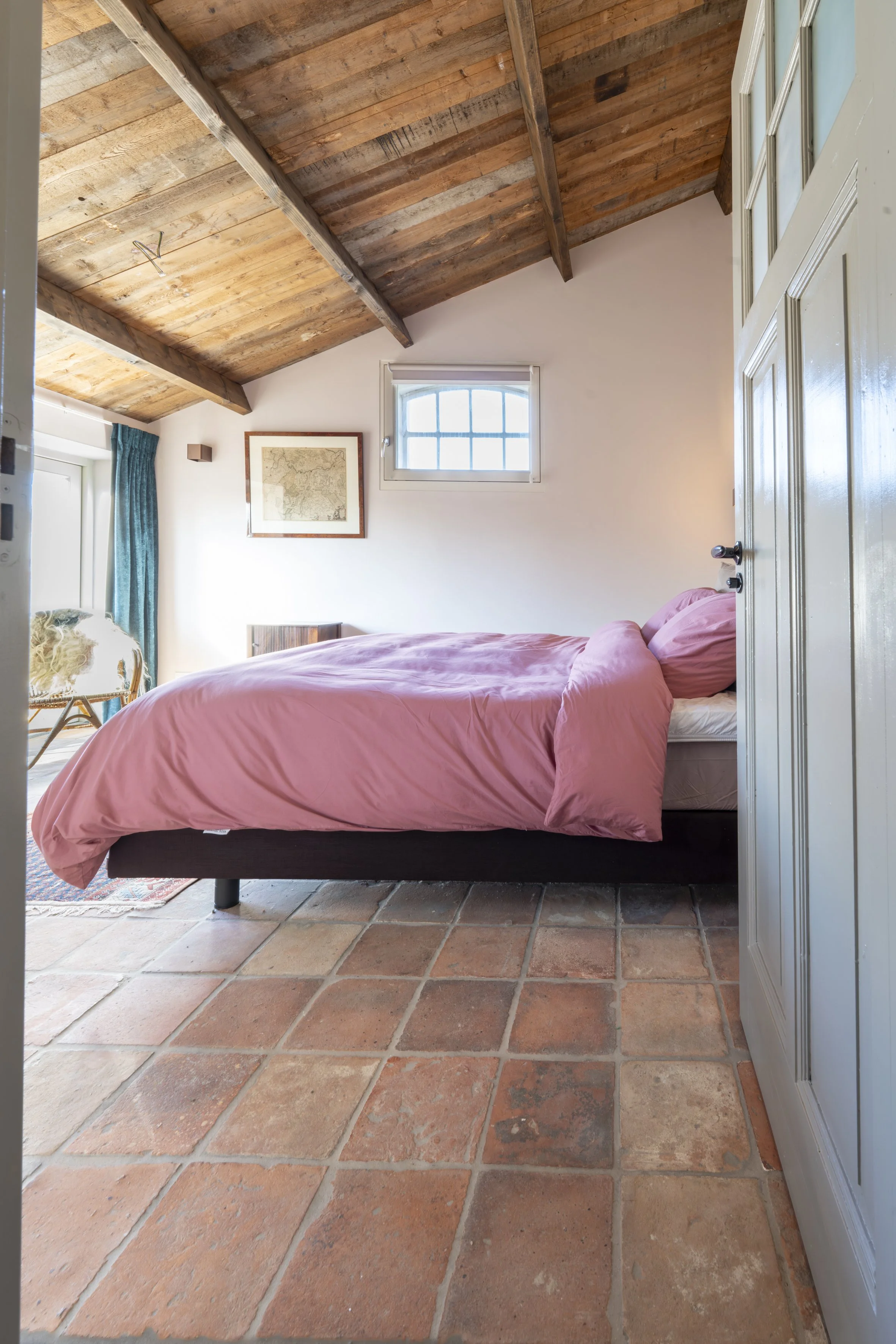 A cozy bedroom with terracotta tile flooring, featuring a bed with pink bedding, a wooden ceiling with exposed beams, a small window, and a framed map on the wall.