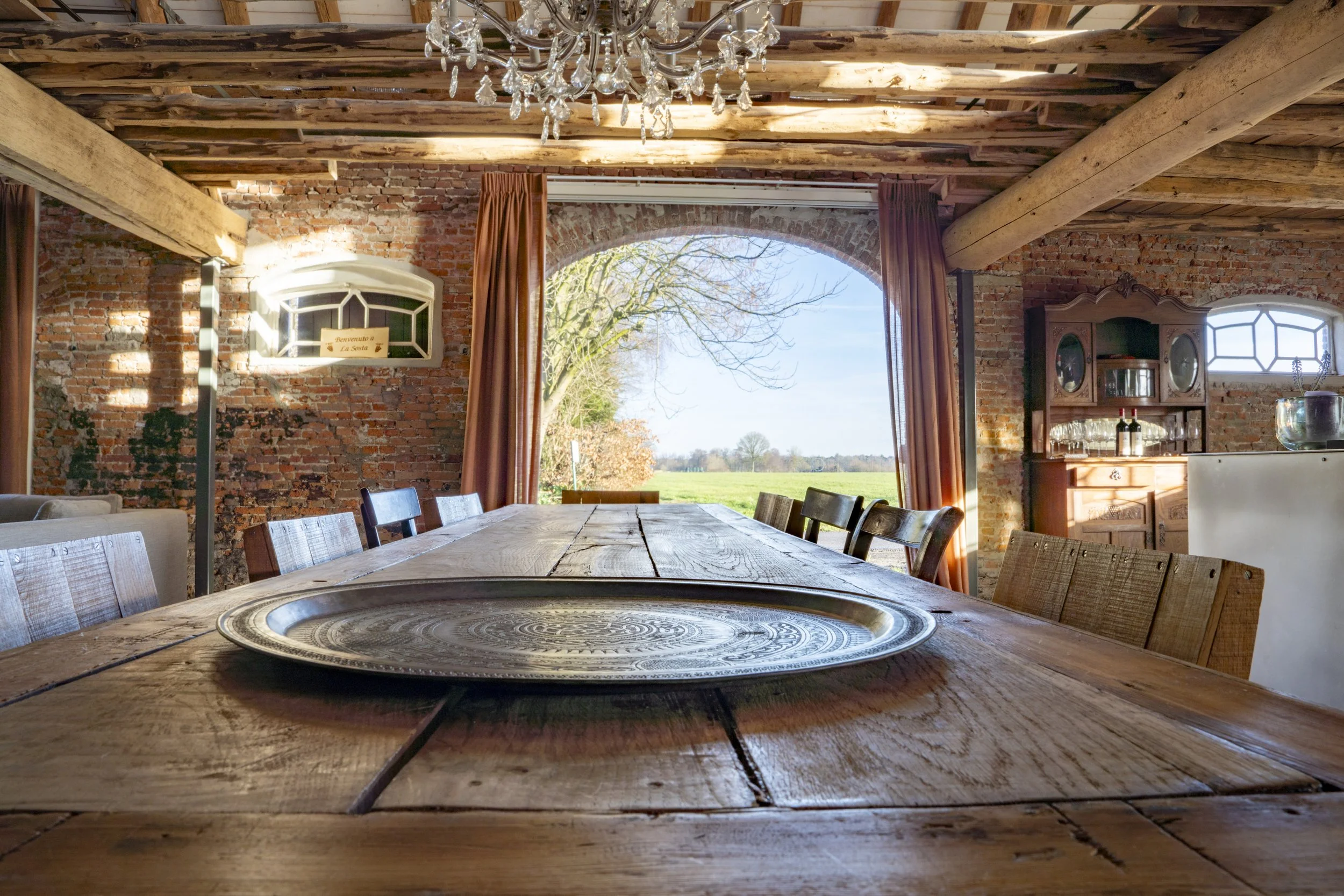 A rustic dining room with brick walls, wooden beams, and a large window showing a view of trees and open fields. The central feature is a long wooden table with chairs, and there is a chandelier hanging from the ceiling.